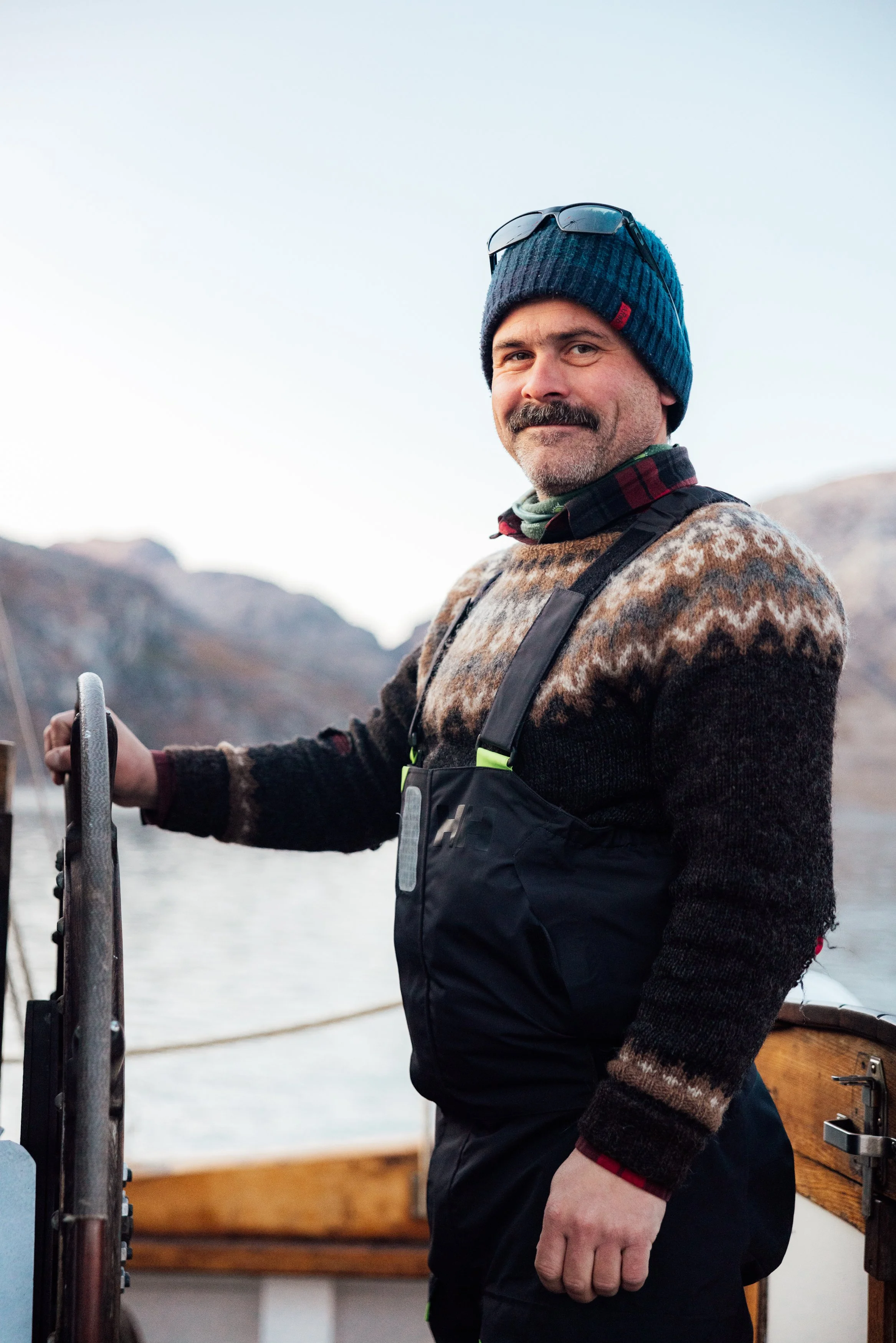 A man in a patterned sweater, beanie, and sunglasses on his head stands on a boat, holding the steering wheel, with mountains and water in the background.