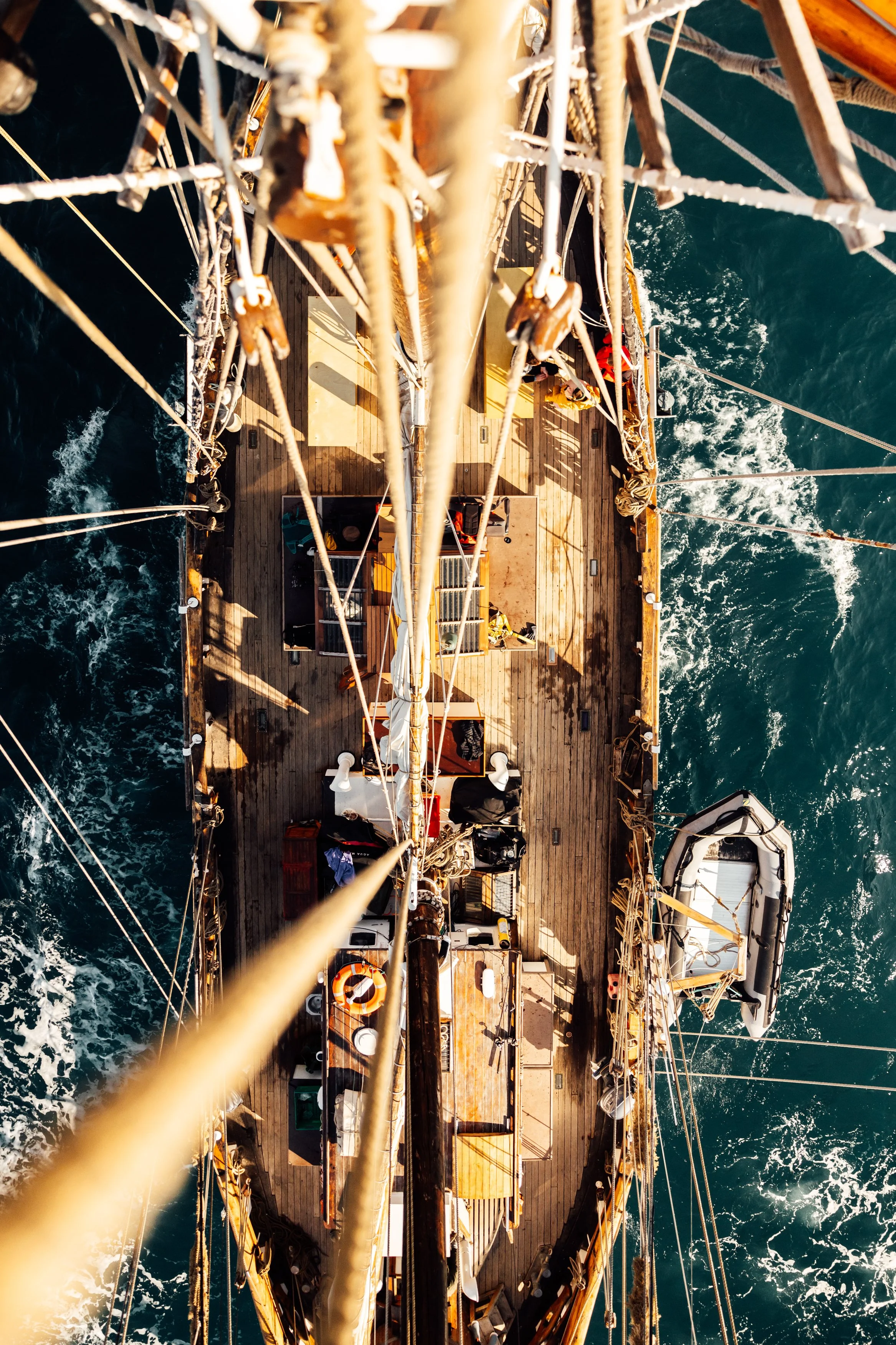 Overhead view of a sailing ship on the water, showing the ship's deck, rigging, and a small inflatable boat attached to the side.