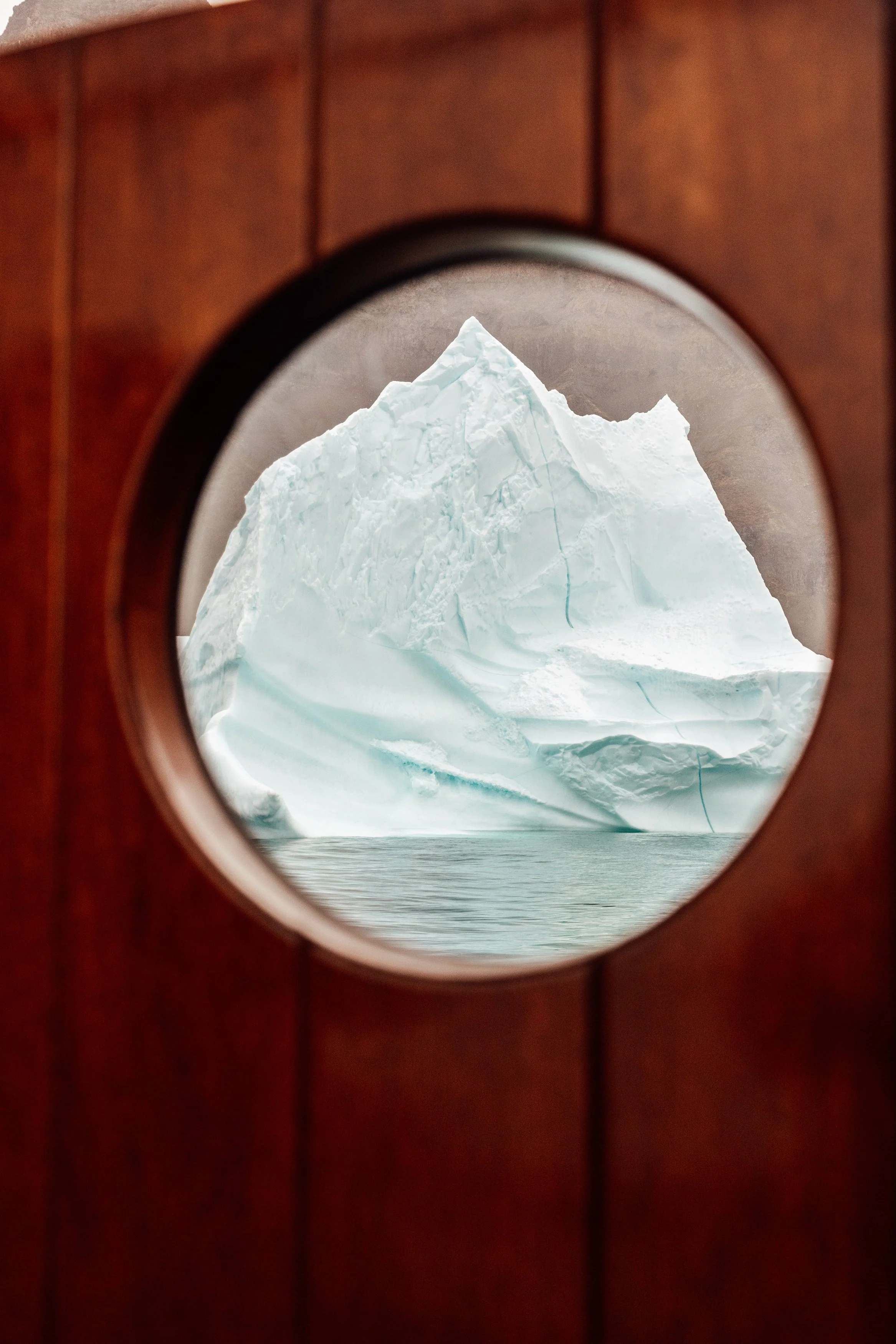 A view of a large iceberg through a circular porthole on a wooden ship or structure.