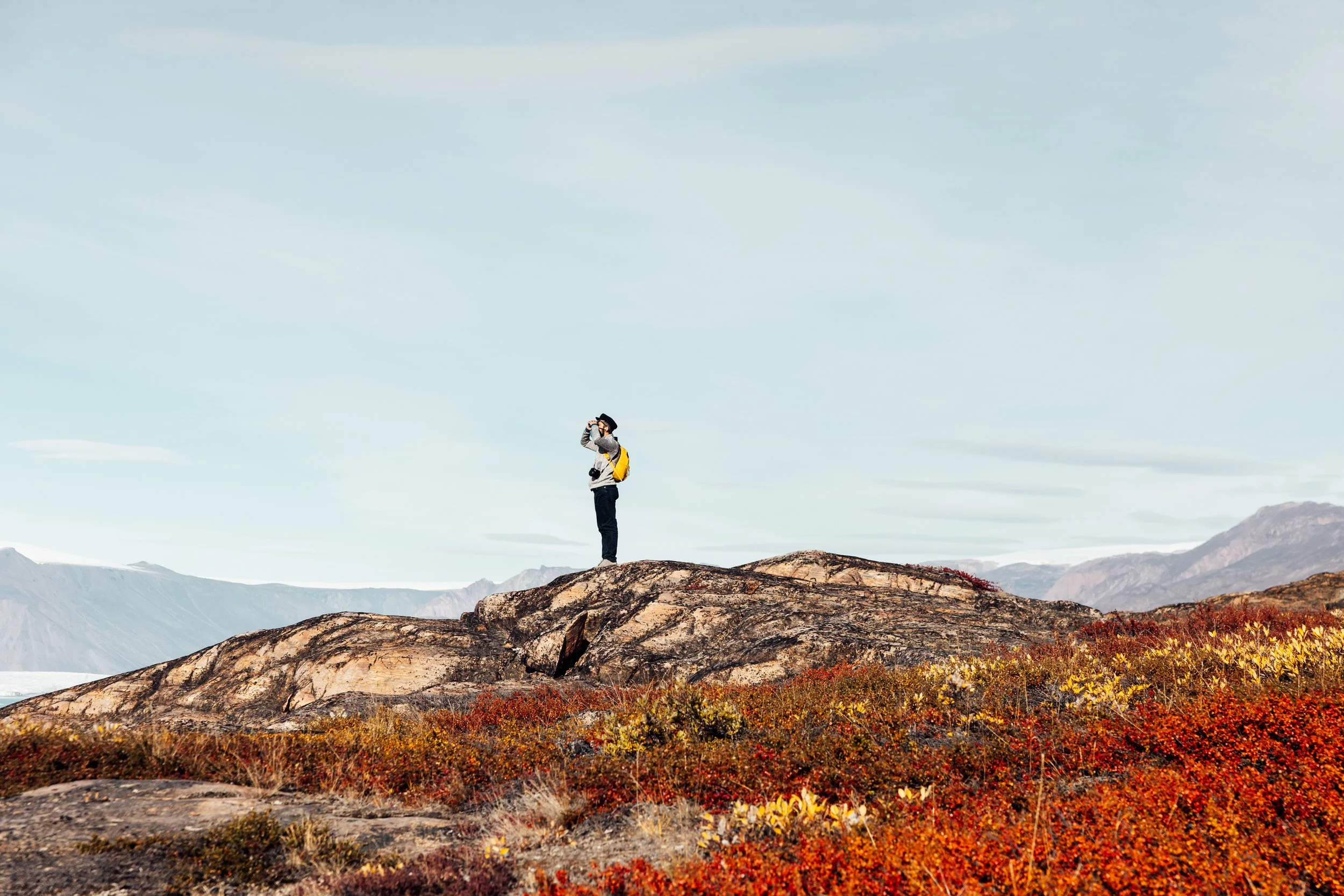 A person standing on a large rock formation during autumn, holding binoculars and looking into the distance, with colorful foliage in the foreground and mountains in the background.