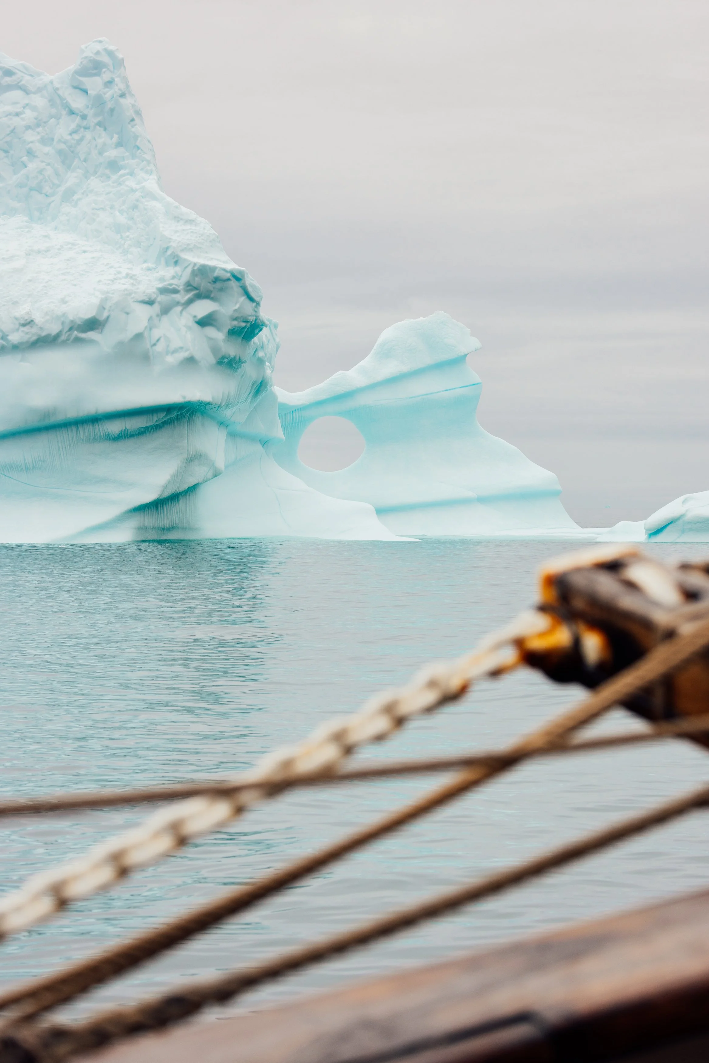 Iceberg with a hole, seen from a boat in cold ocean water, overcast sky.