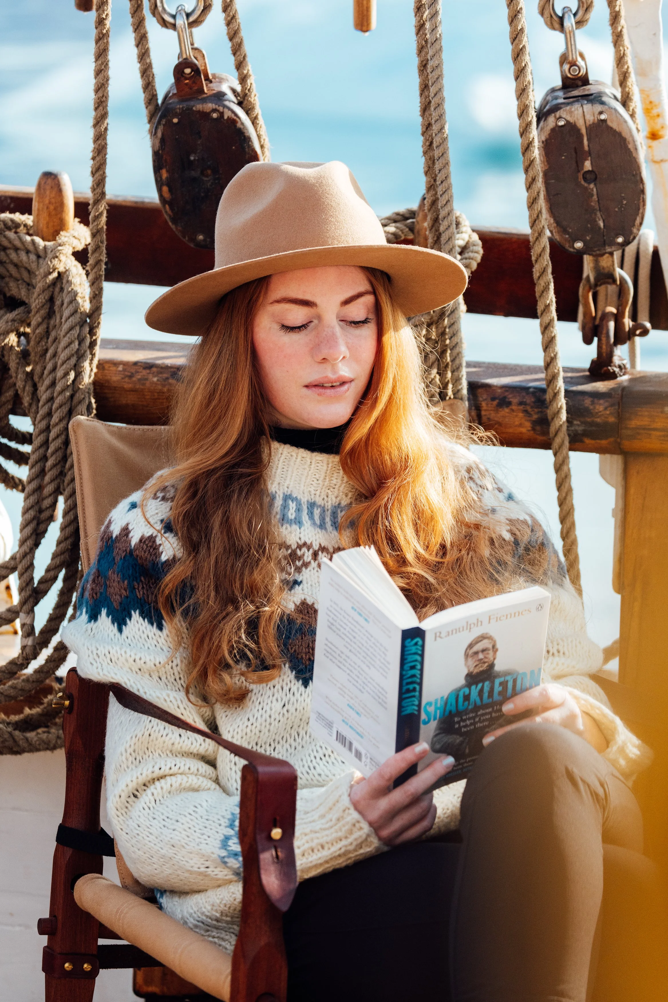 A woman with long red hair, wearing a beige wide-brimmed hat and a cream-colored sweater with a blue and brown pattern, is sitting and reading a book titled 'Shackleton' on a boat with nautical ropes and wooden structures, and water in the background