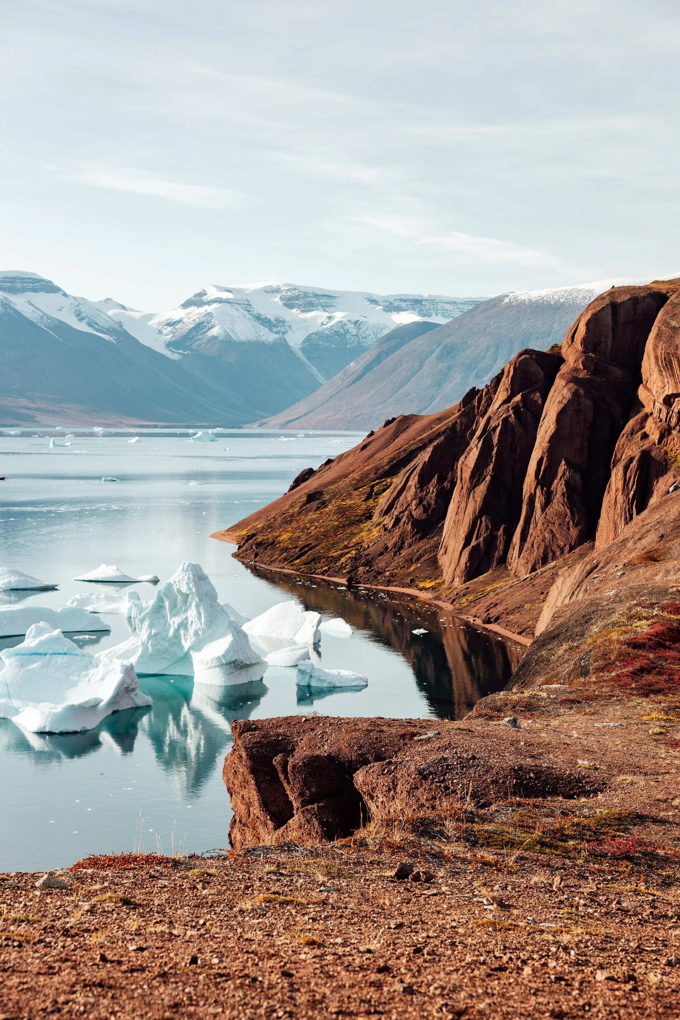Scenic landscape of a glacial lake with floating icebergs, surrounded by reddish-brown rocky cliffs and snow-capped mountains in the background.