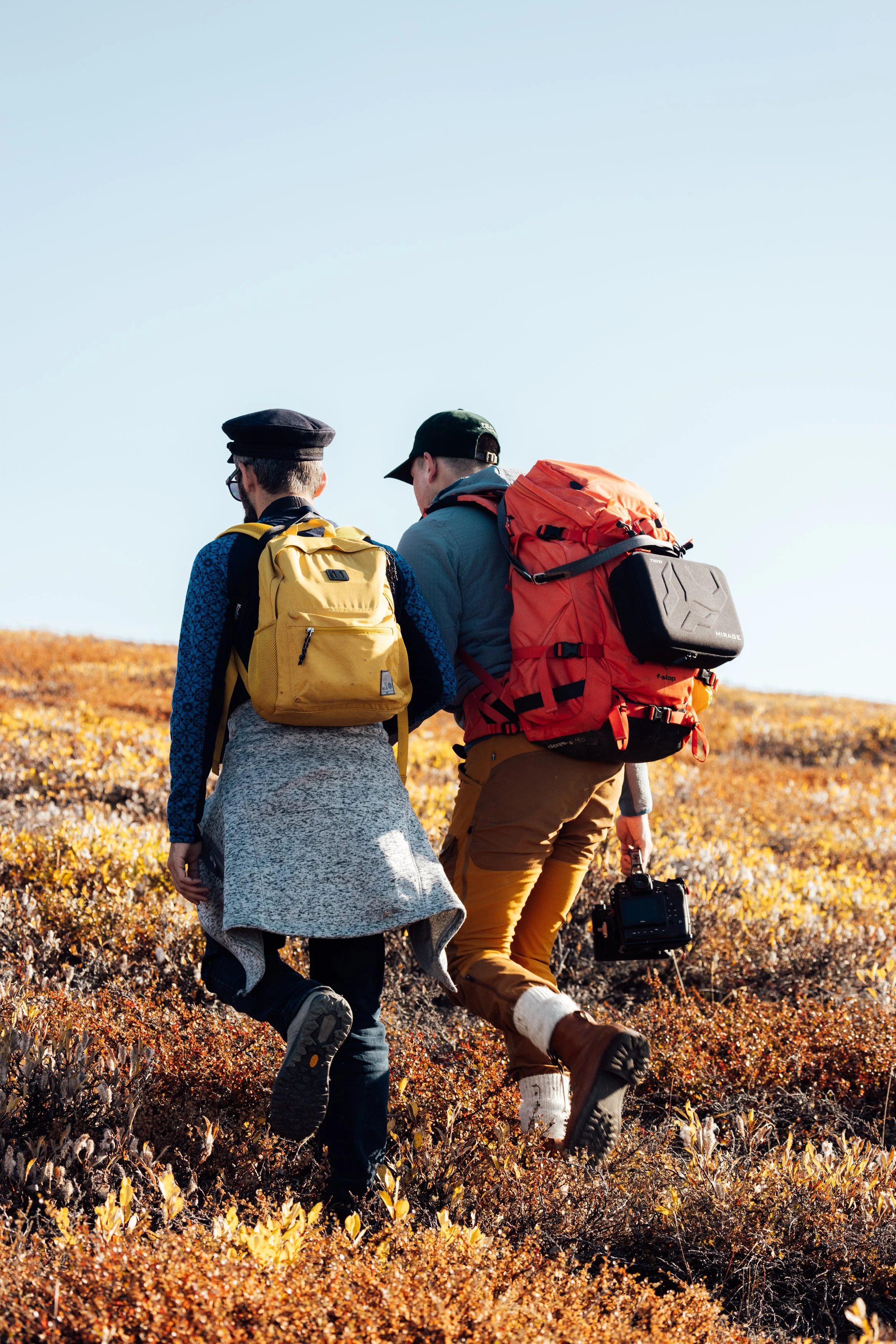 Two people hiking through a grassy, autumn-colored landscape with backpacks, carrying a camera, under a clear blue sky.