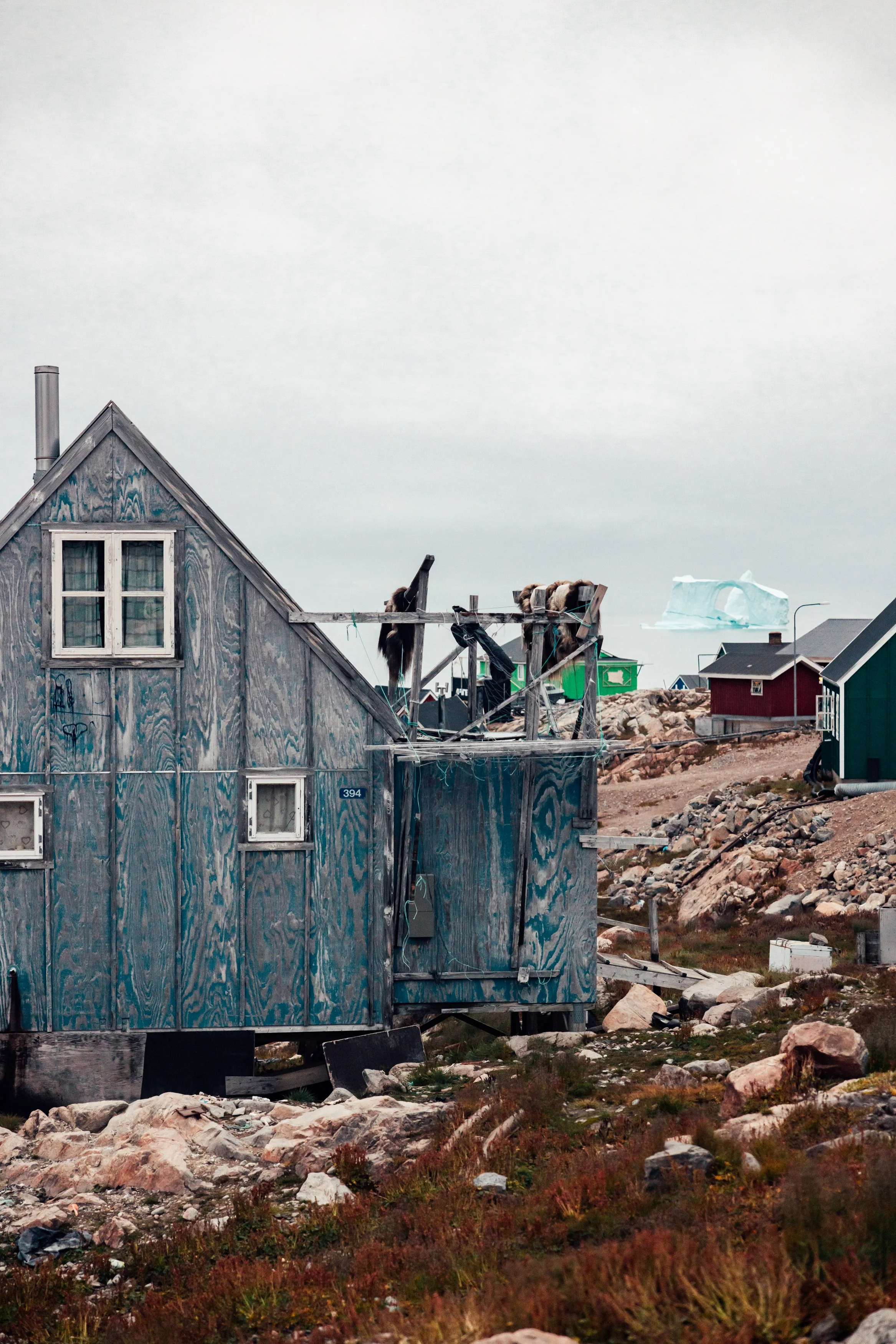 A weathered wooden house with a damaged roof under construction, surrounded by rocky terrain and other small houses in the background, with an iceberg floating in the sky.