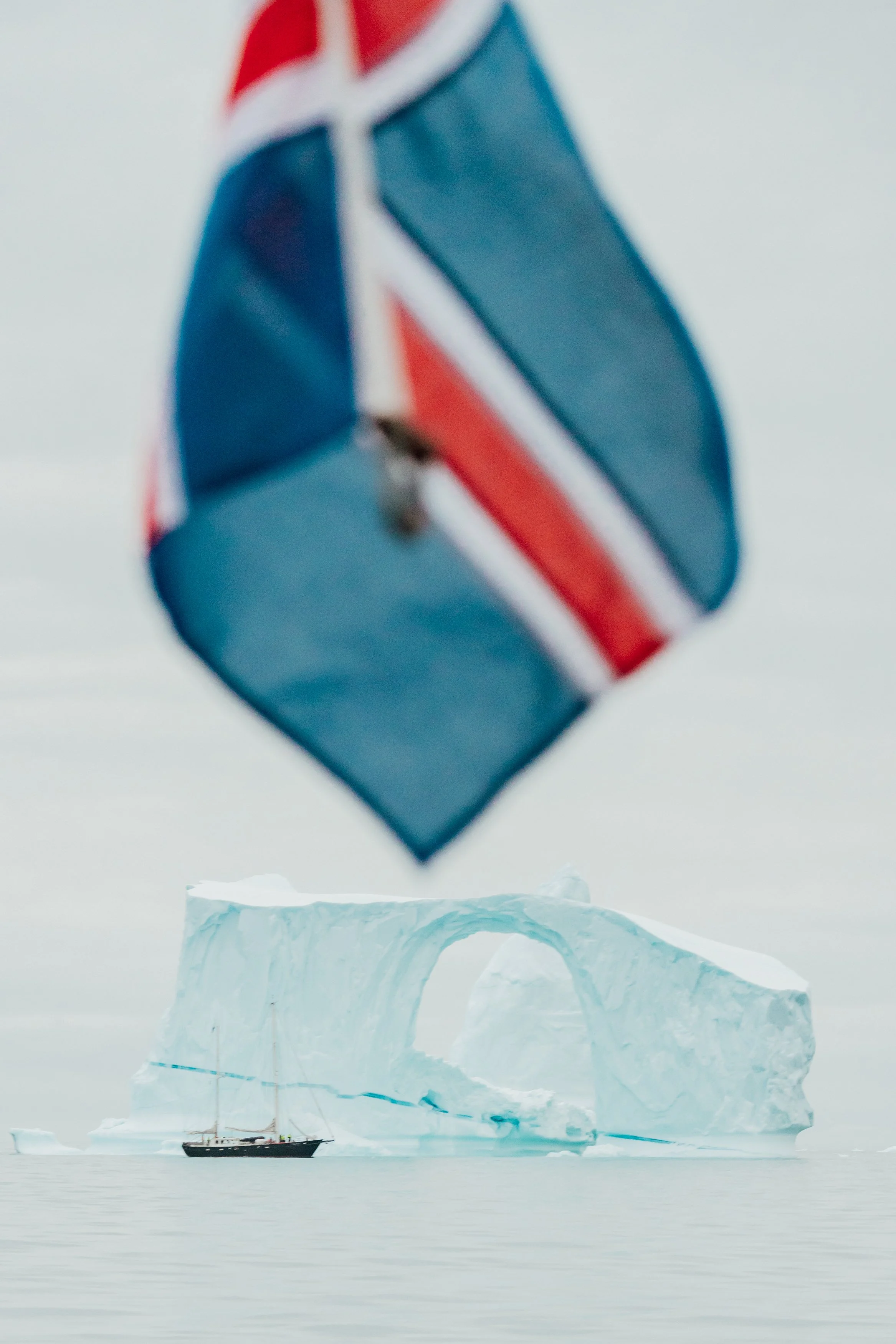 A small sailboat sailing near a large iceberg with an arch in it, viewed from below a drones with a Union Jack design in the foreground.