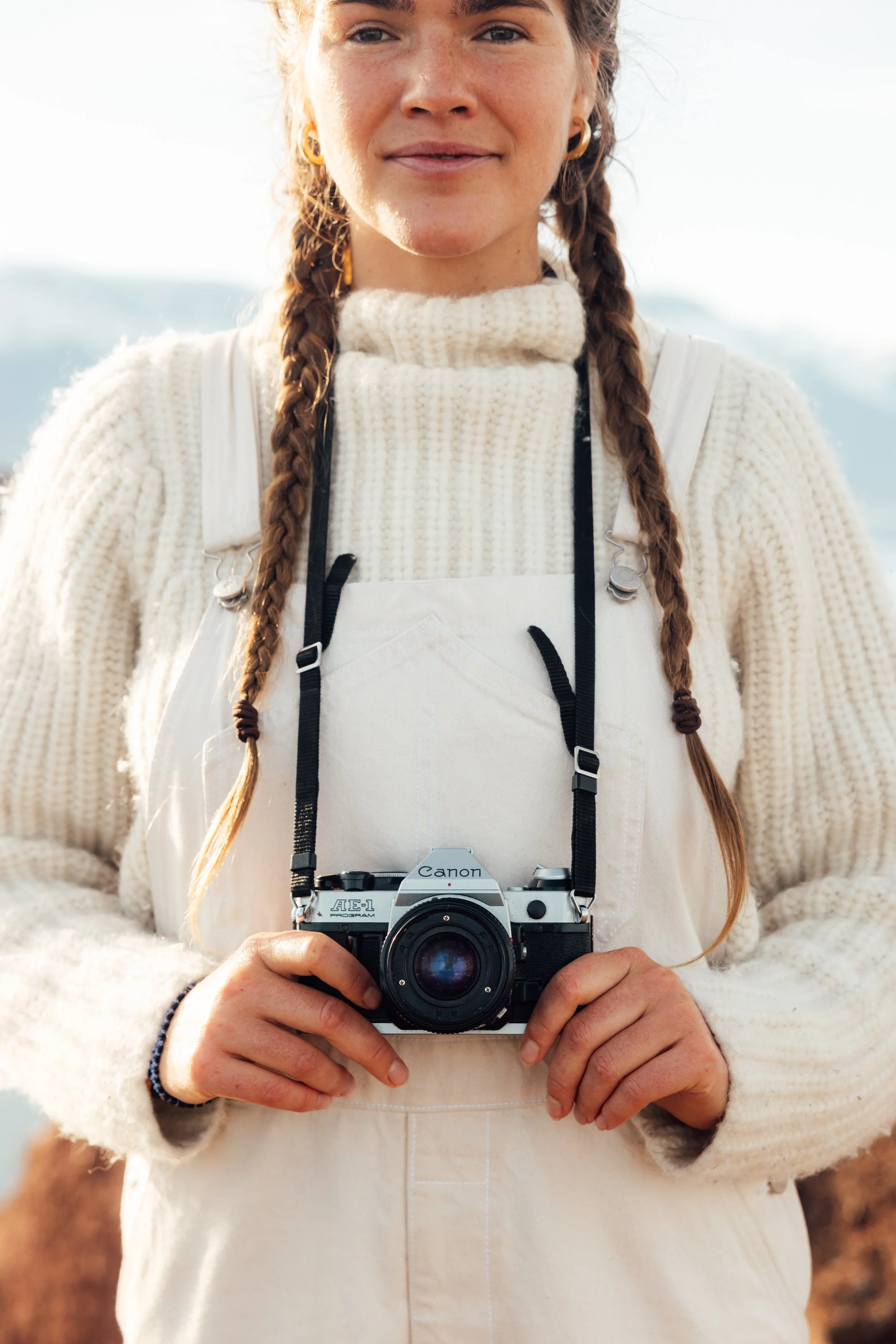 A woman with braids wearing a cream sweater and overalls holding a vintage film camera outdoors.