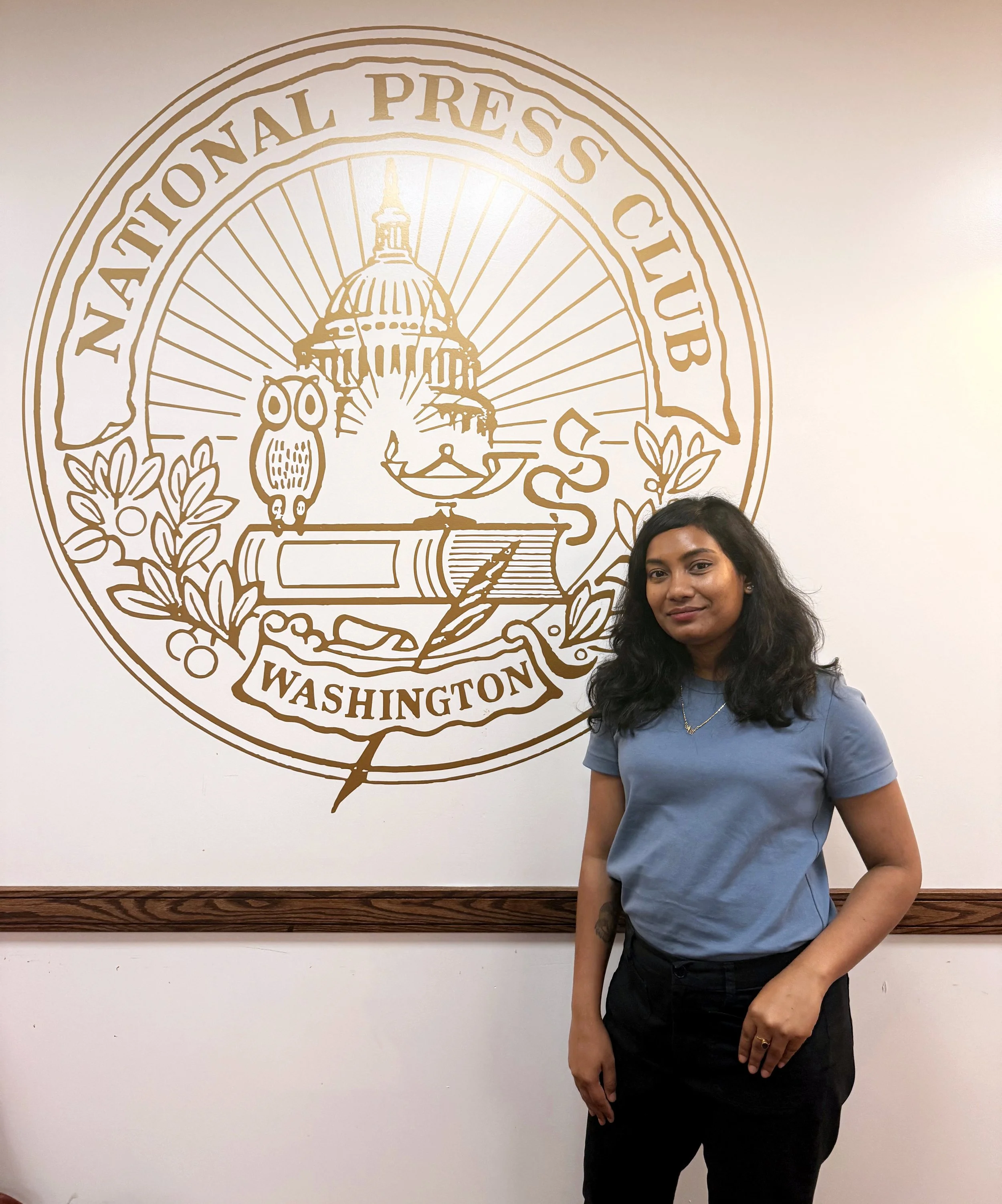 A woman with long dark hair, wearing a blue t-shirt and black pants, standing in front of a wall with the emblem of the National Press Club of Washington. The woman is smiling slightly, with her left arm resting at her side and her right hand on her waist.