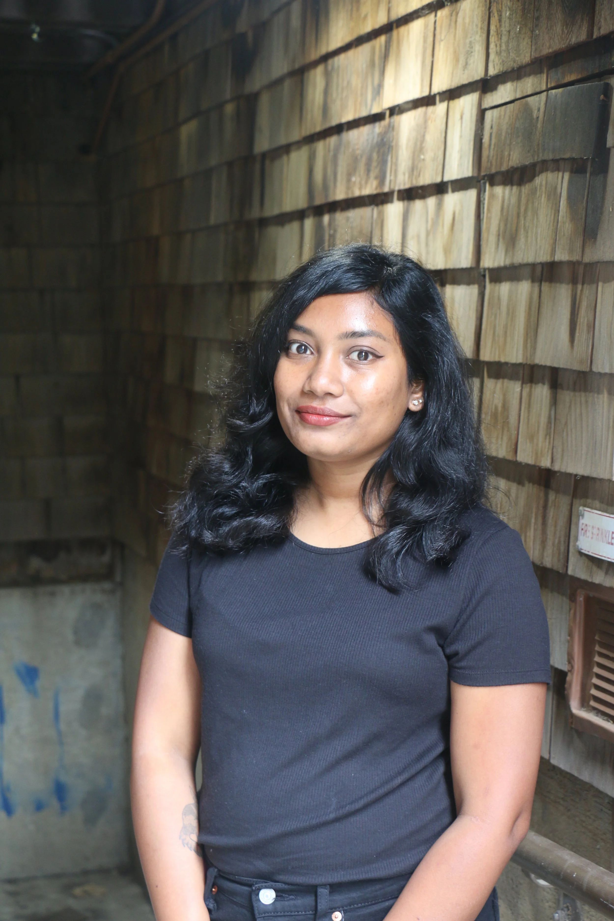 A woman with black wavy hair, wearing a black shirt and earrings, standing in front of a wooden wall in an indoor setting.
