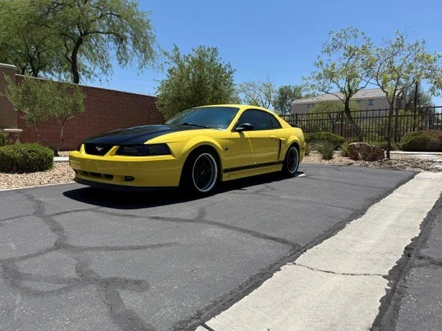 Yellow and black sports car parked on a paved driveway next to a sidewalk with desert landscaping and trees in the background.