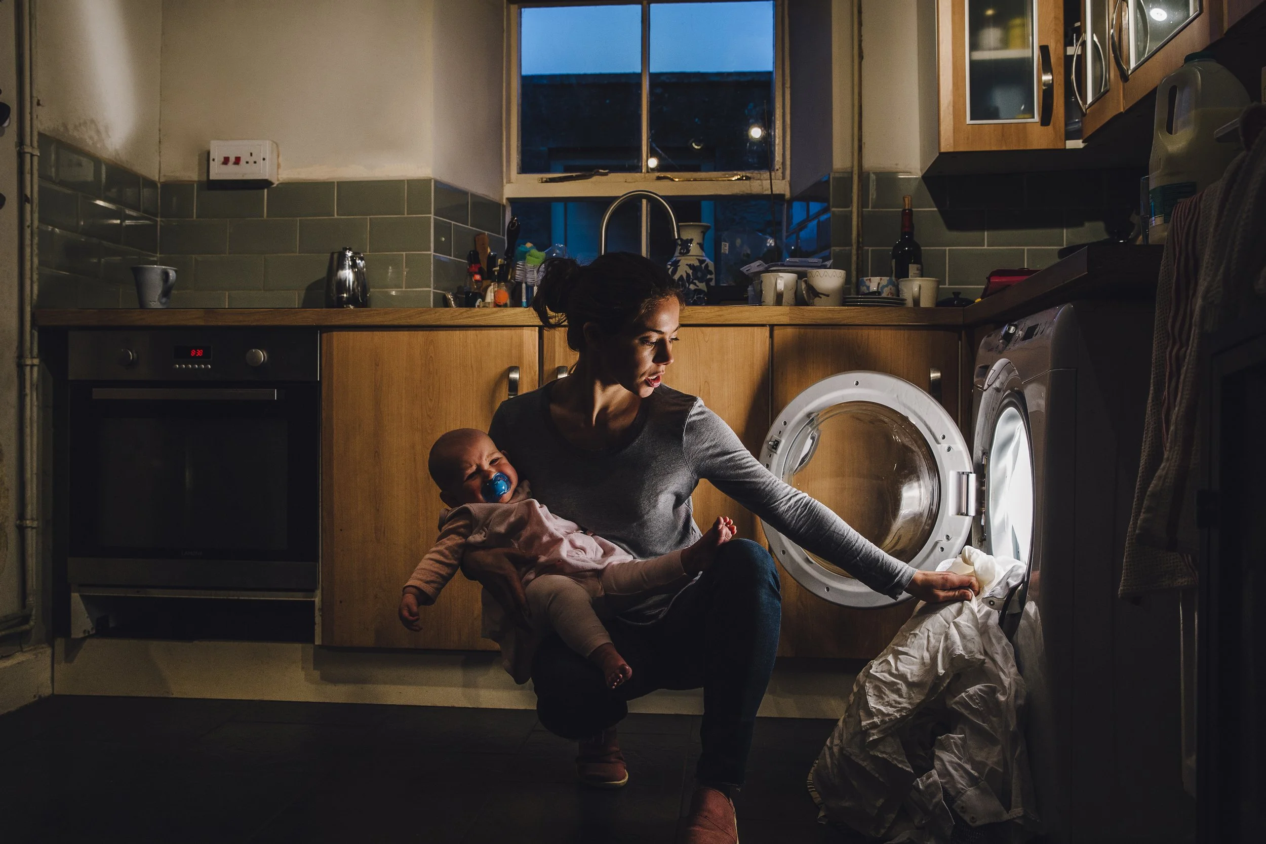 A woman with dark hair tied back, sitting on the floor in a kitchen, holding a crying baby with pacifier, as she looks at the washing machine door. The kitchen has wooden cabinets, a window showing evening sky, and various kitchen items on countertops.