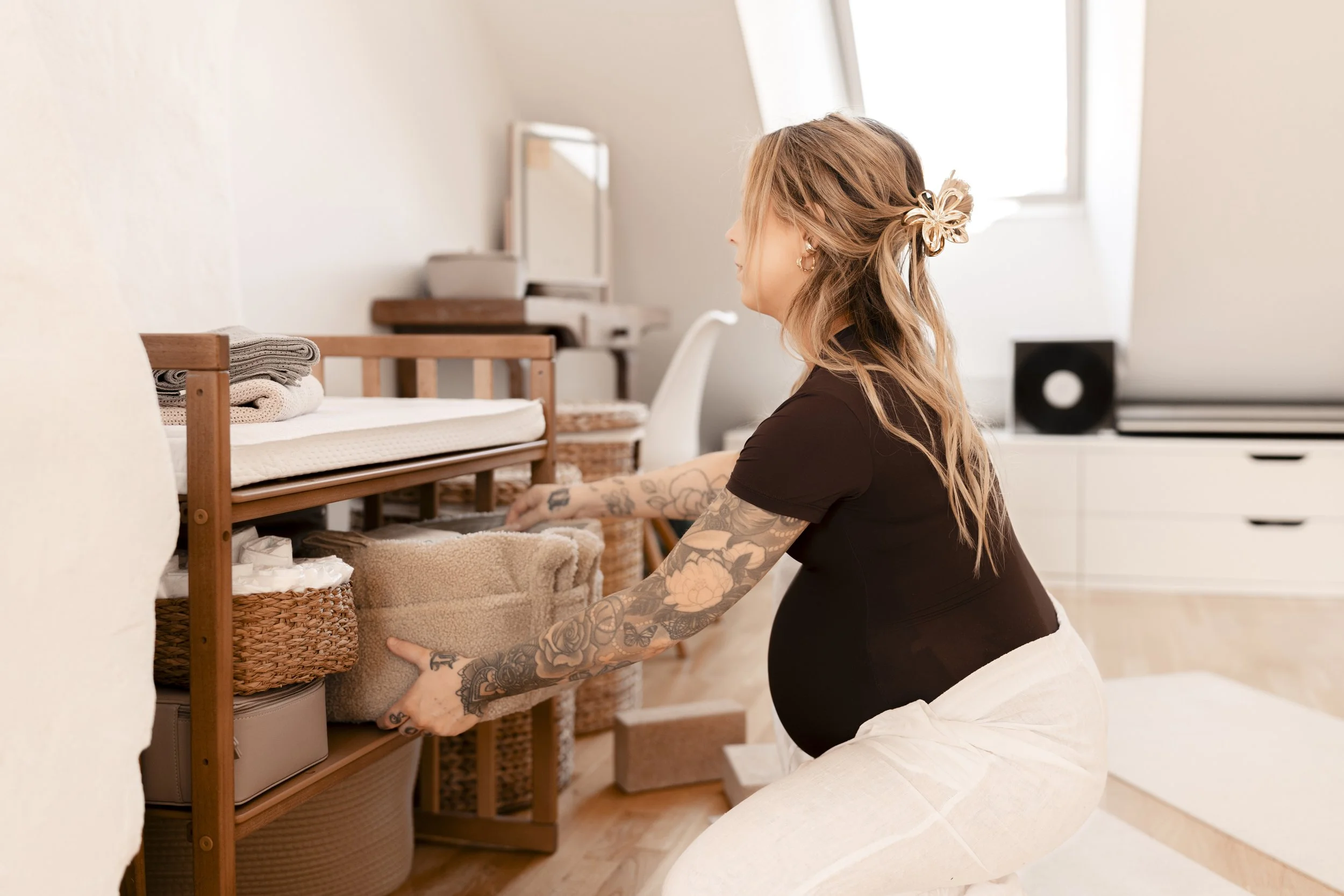 A woman with tattoos on her arms wearing a black shirt and white pants is kneeling and organizing baskets filled with folded towels on a wooden shelf in a bright room with white walls and windows.