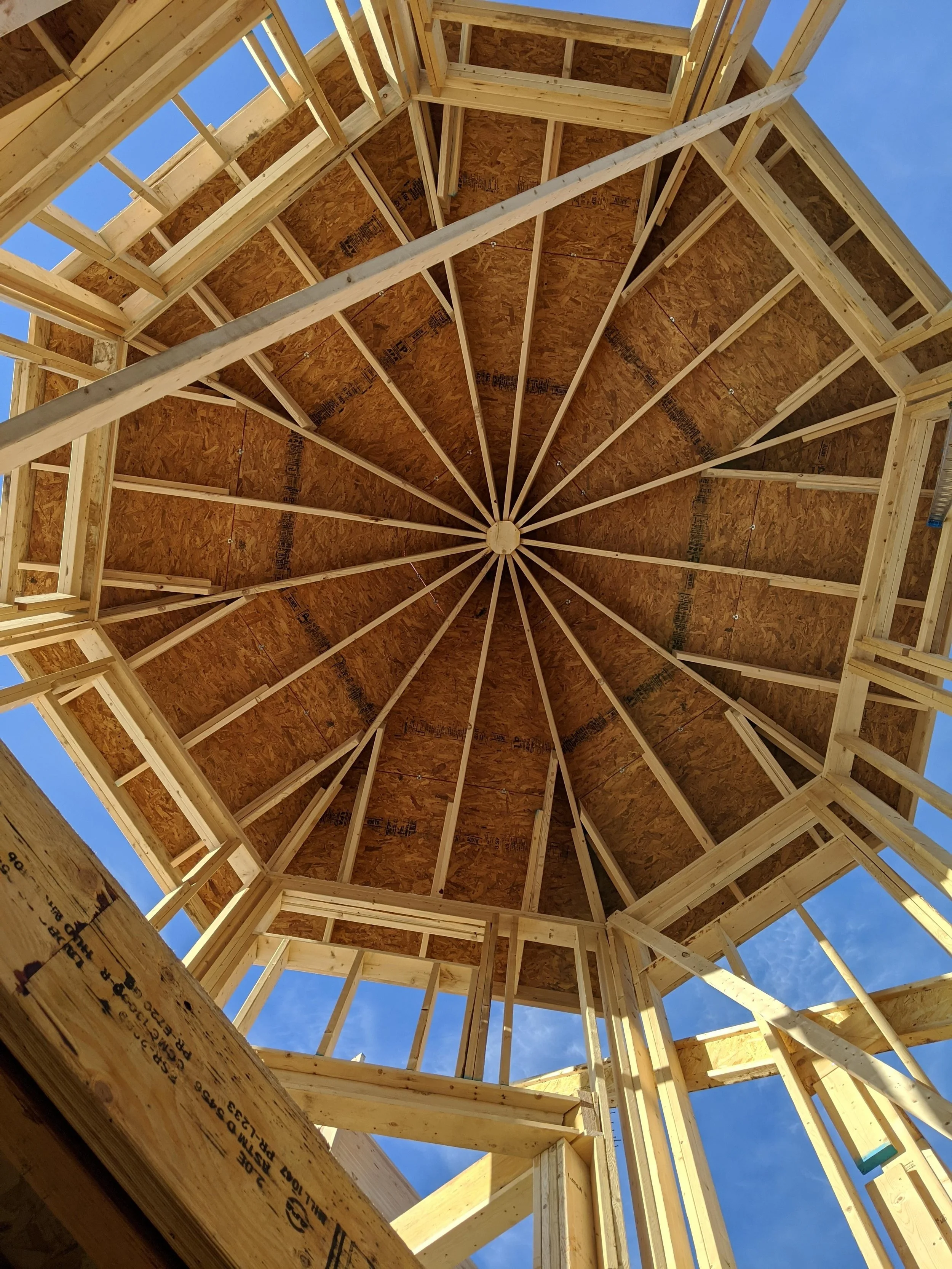 View looking up at the wooden framework of a building under construction, showing roof trusses and framing against a blue sky.