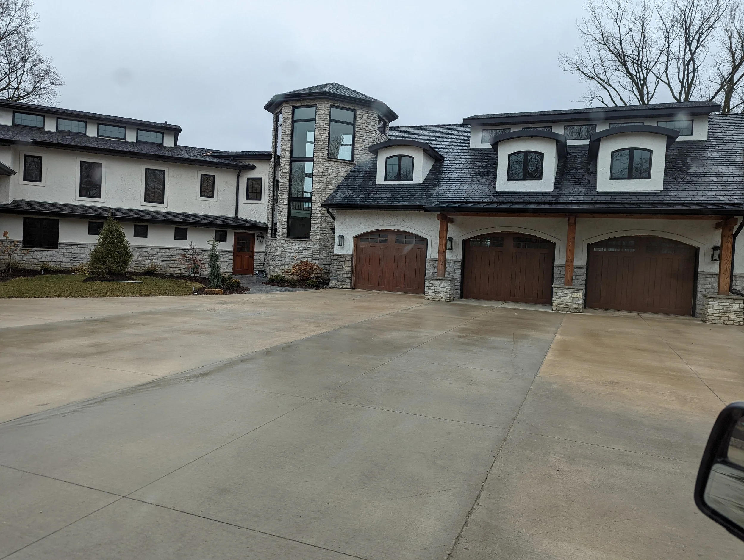 Large modern house with multiple windows, stone and stucco exterior, a double garage, and a small landscaped front yard with plants and trees, on a cloudy day.