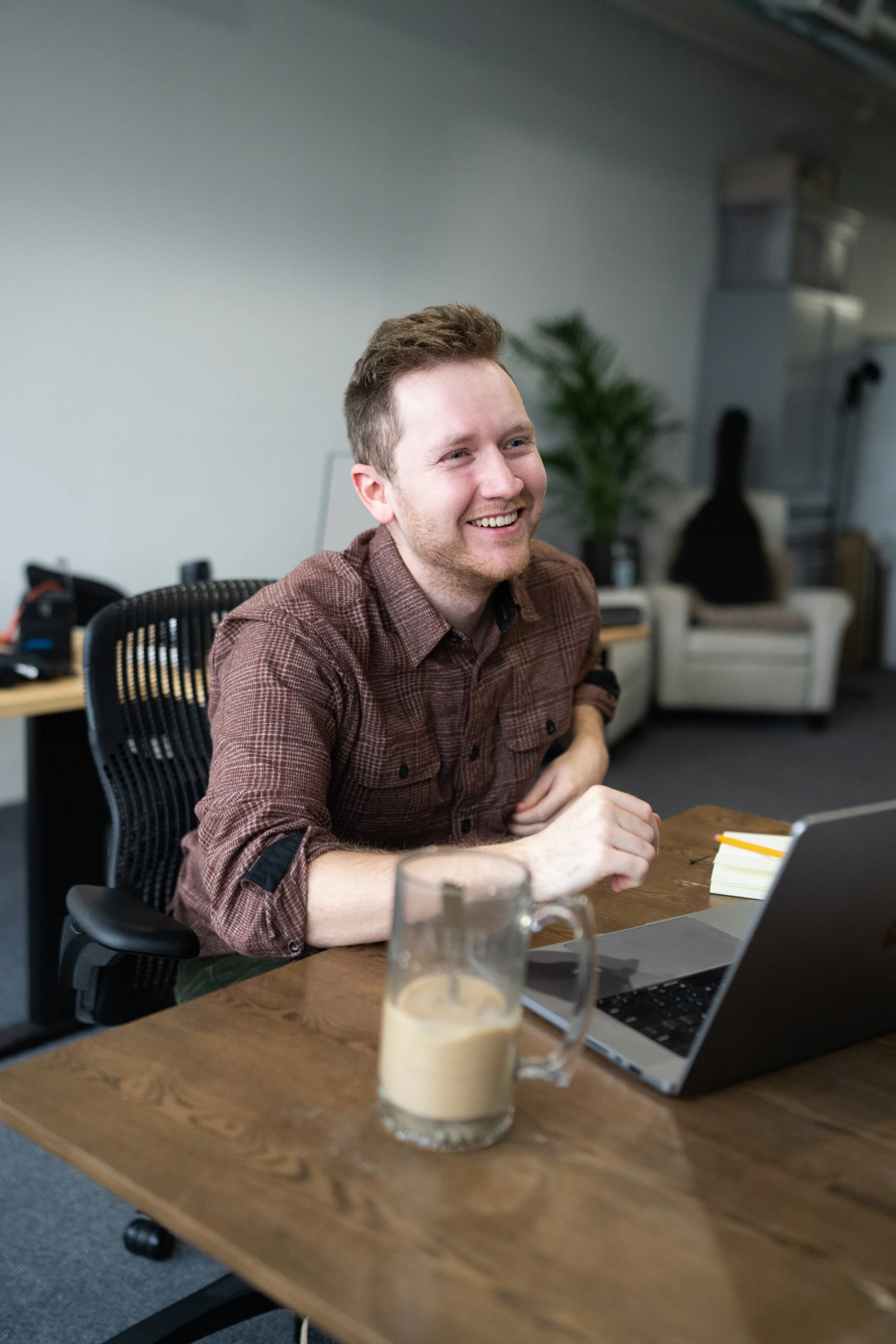 A young man with short brown hair and a beard sitting at a wooden desk in an office, smiling and looking to his right. He is wearing a brown checkered shirt. On the desk in front of him are a laptop, a notebook, a yellow pen, and a glass mug with a beverage. In the background, there is a black chair, a large potted plant, and shelves with storage boxes.