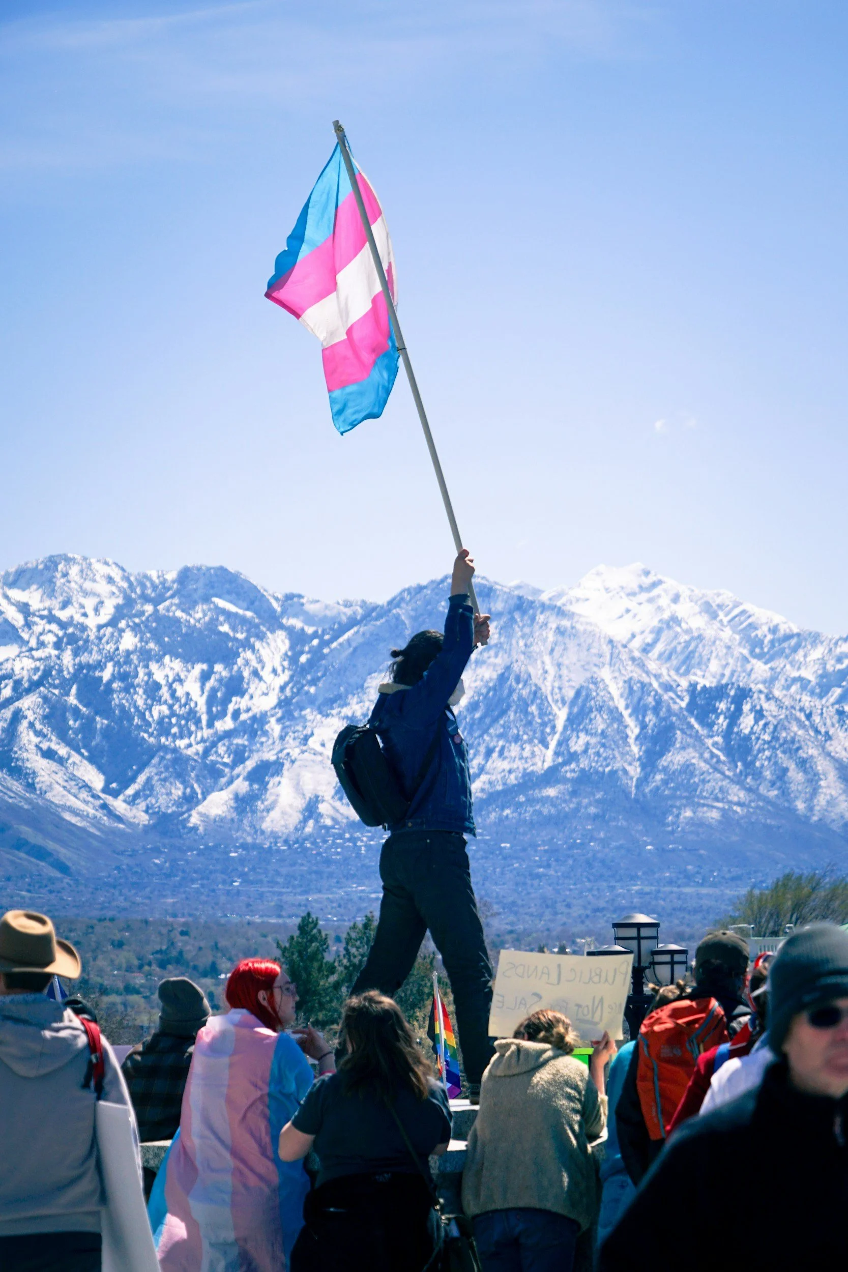 A person holding a transgender pride flag with mountains in the background during a protest or rally, with a crowd of people around.