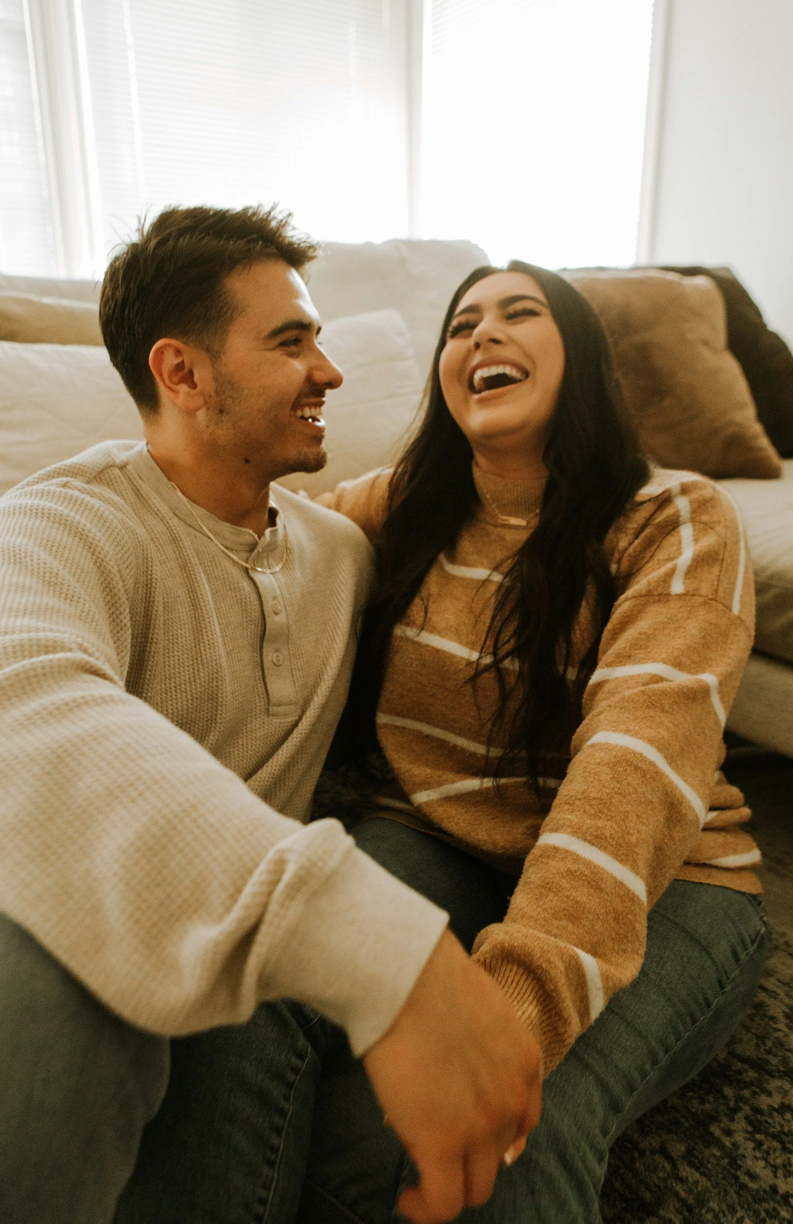 A man and woman sitting together on a sofa, laughing and enjoying each other's company in a cozy living room with abundant natural light.