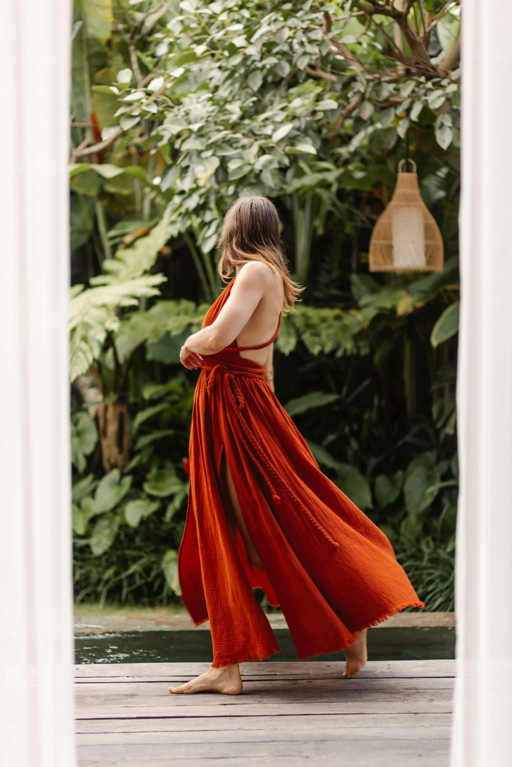 A woman with long brown hair standing on a wooden deck, looking away, wearing an orange-red dress, with lush green foliage and a wicker lantern hanging in the background.