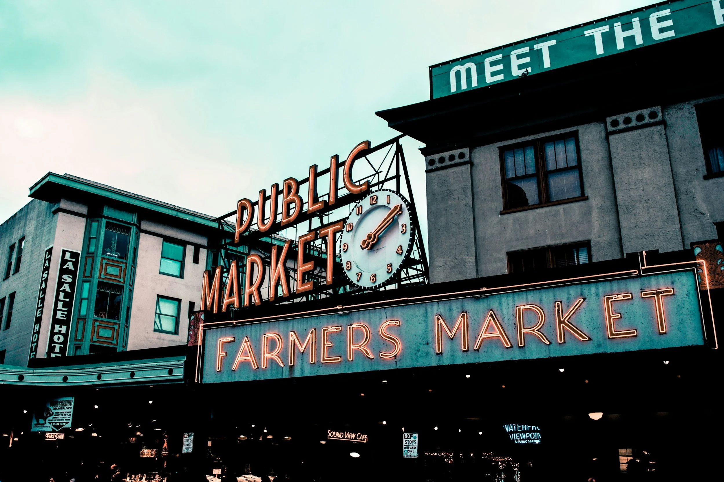 A large vintage-style neon sign that reads 'FARMERS MARKET' with a clock above it displaying the time as 4:10. The sign is mounted on a building facade in an urban setting, with other buildings and signs visible in the surroundings.