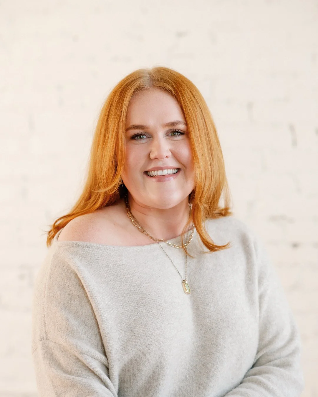 Portrait of a woman with red hair smiling, wearing a light beige sweater and layered necklaces, against a white brick wall.