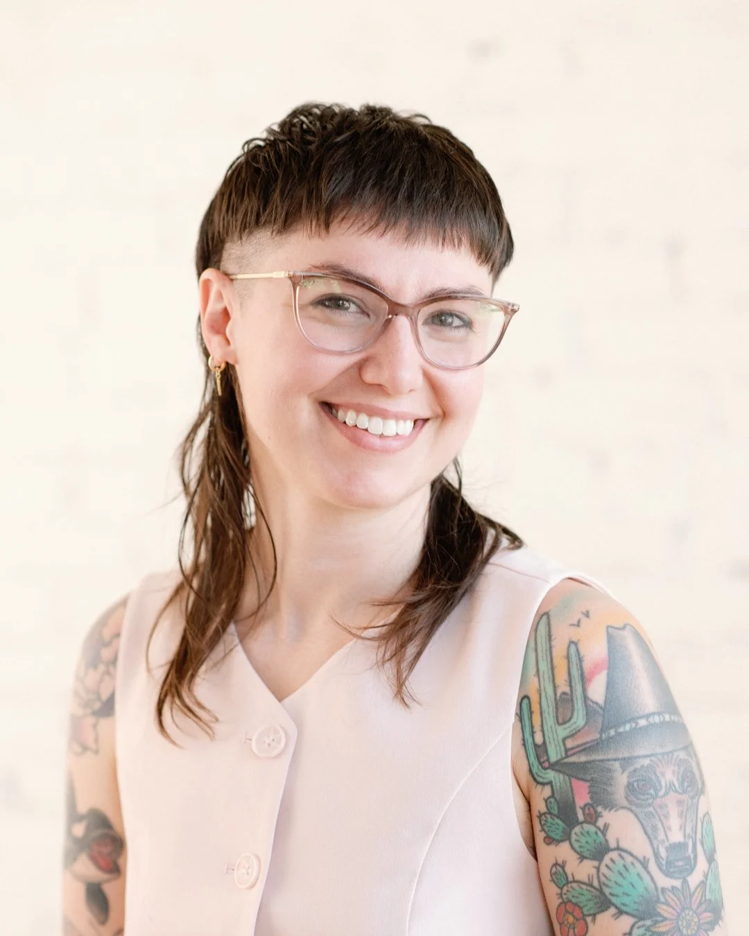 A woman with short brown hair, glasses, and tattoos on her arms, smiling and wearing a light-colored sleeveless top.