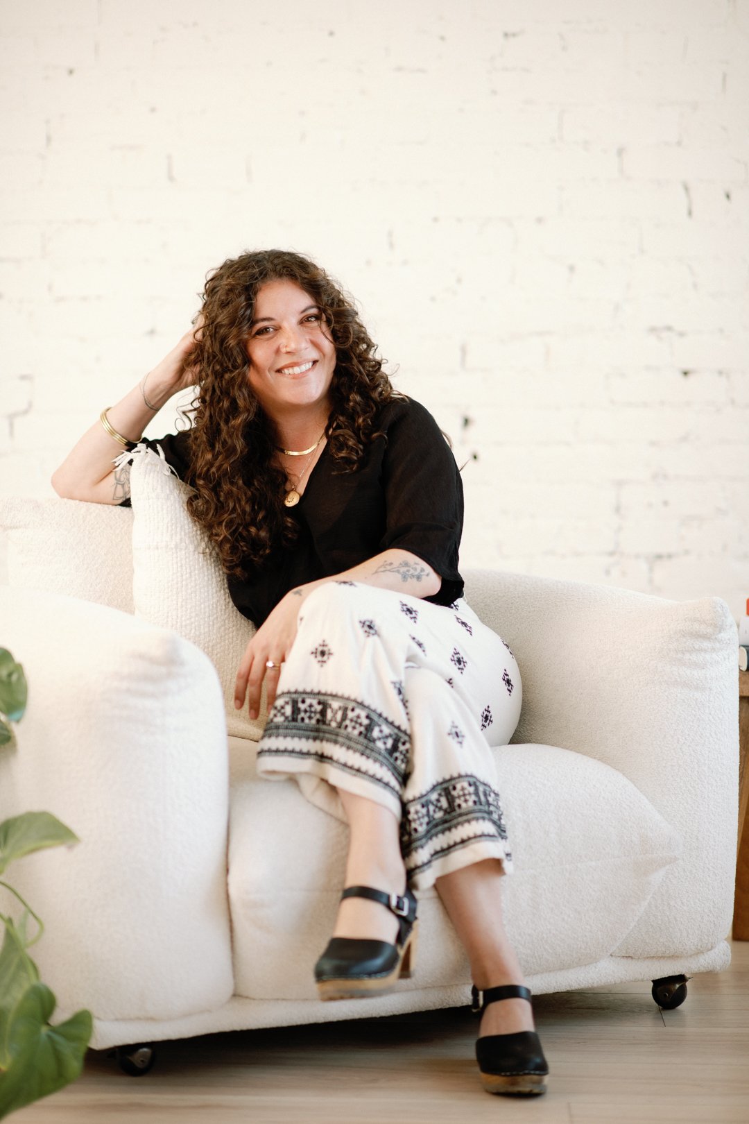 A woman with curly hair sitting on a white couch, wearing a black top, patterned white pants, and black heels, smiling at the camera against a white brick wall background.