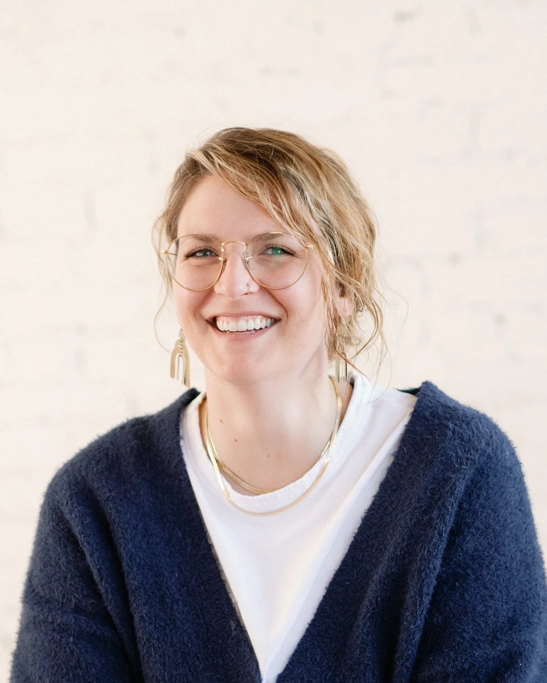 A woman with short, curly blonde hair and glasses smiling against a white wall, wearing a navy blue cardigan, white t-shirt, and gold jewelry.
