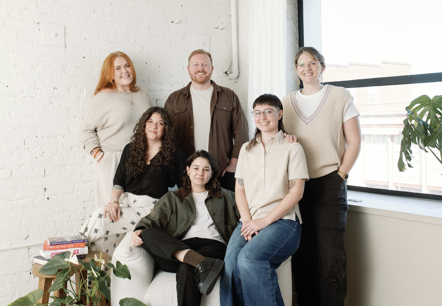 A diverse group of seven people, including men and women of various ethnicities, smiling and posing together in a brightly lit room with a white brick wall and large window, with plants and books in the foreground.