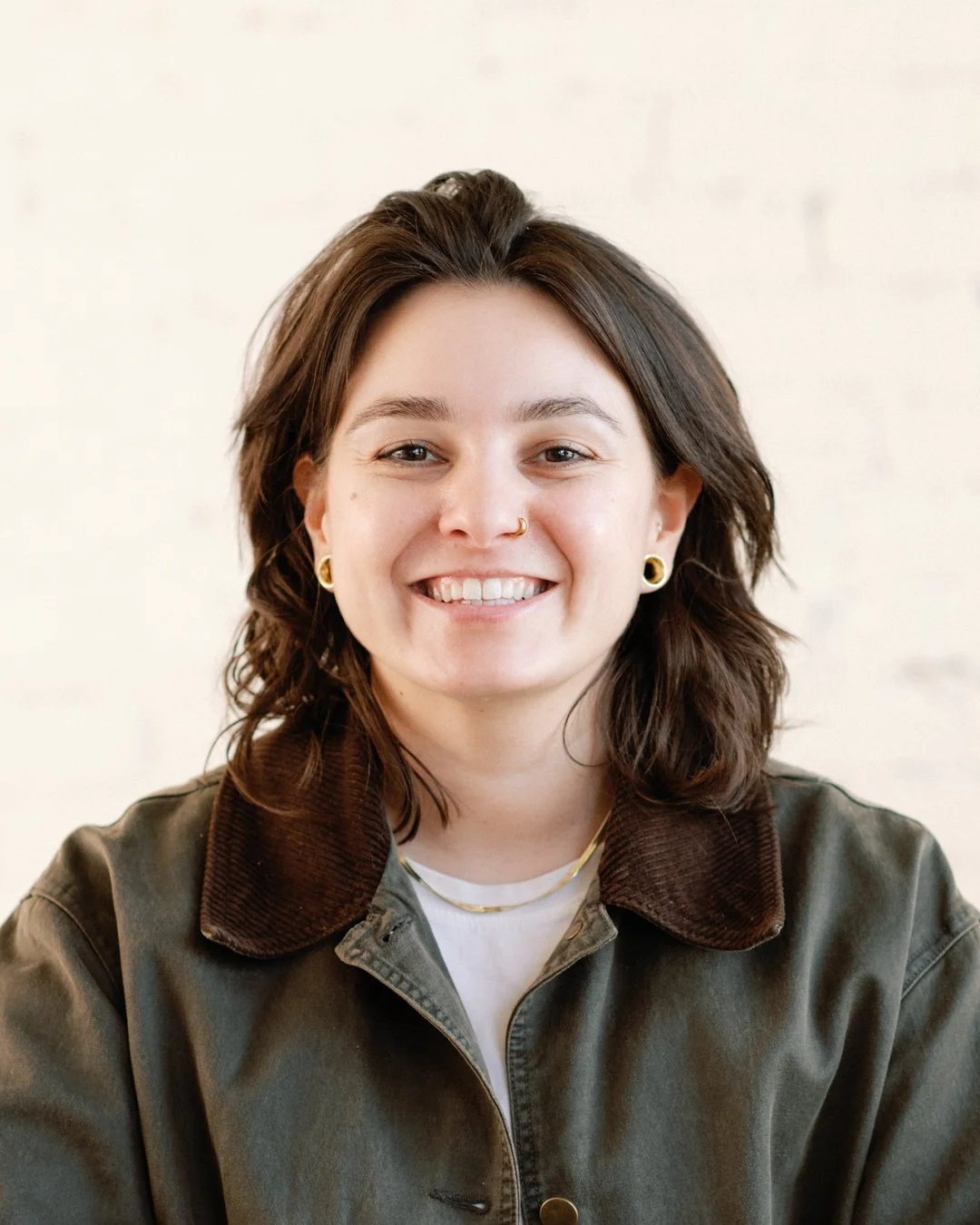 A young woman with shoulder-length dark brown hair, wearing a green jacket with a brown collar, gold earrings, and a gold necklace, smiling against a plain light background.