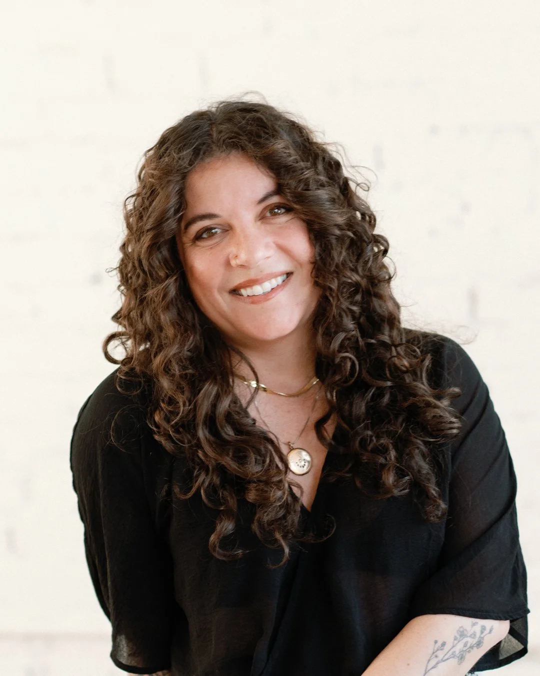 A woman with long, curly brown hair wearing a black top and a gold necklace, smiling at the camera against a light background.