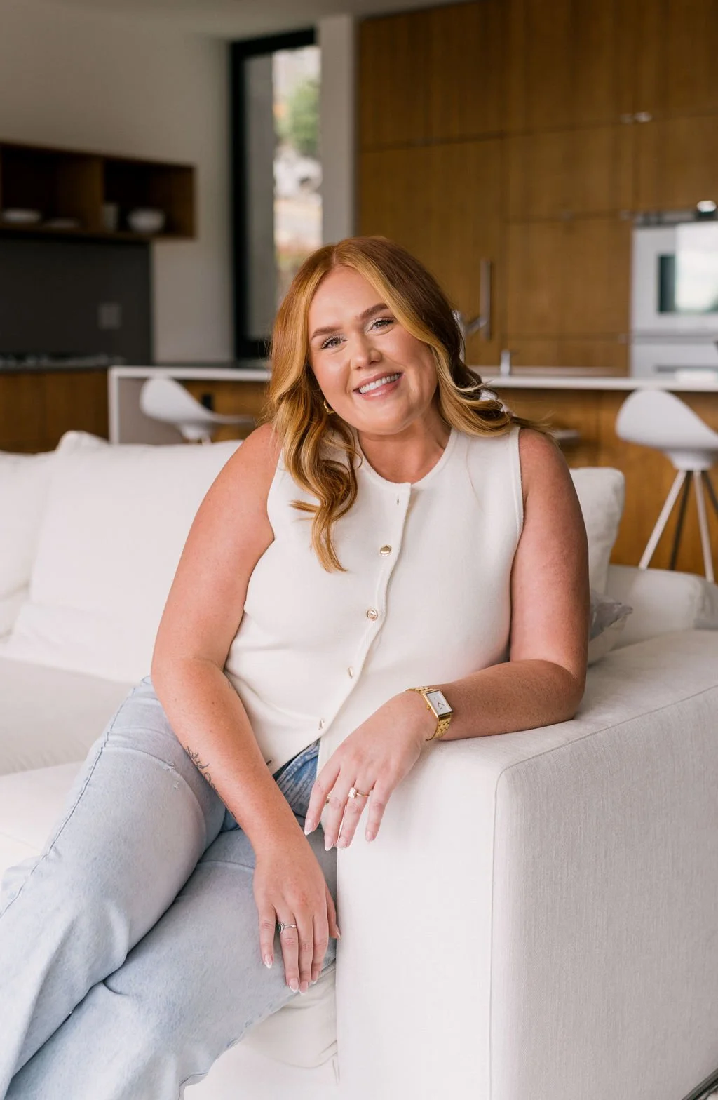 A smiling woman with red hair sitting on a white sofa in a modern kitchen with wooden cabinets, a window, and a built-in microwave.