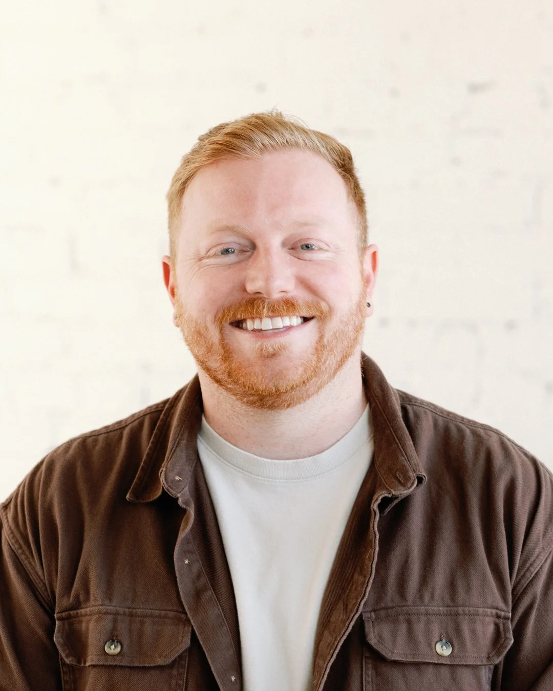 A smiling man with red hair and beard, wearing a brown jacket over a white shirt, standing against a white wall.