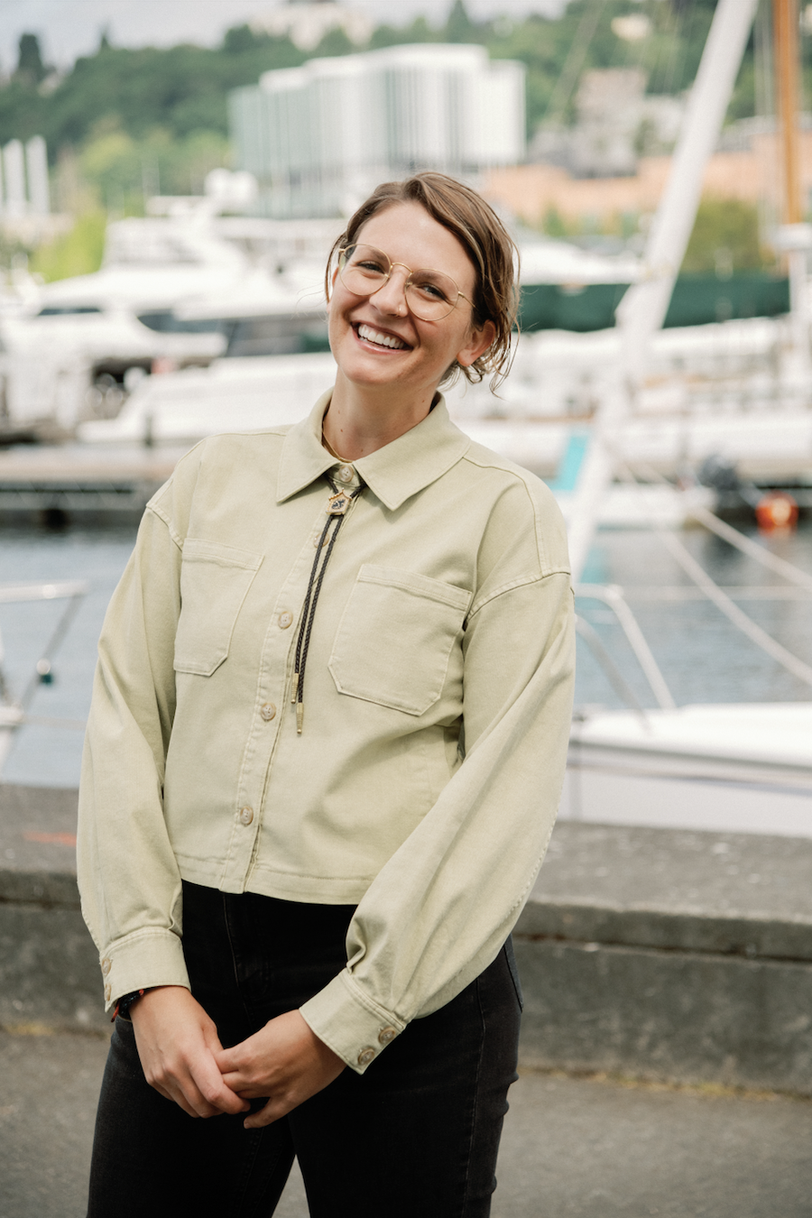 A smiling woman with short brown hair and glasses standing in front of a marina with boats.