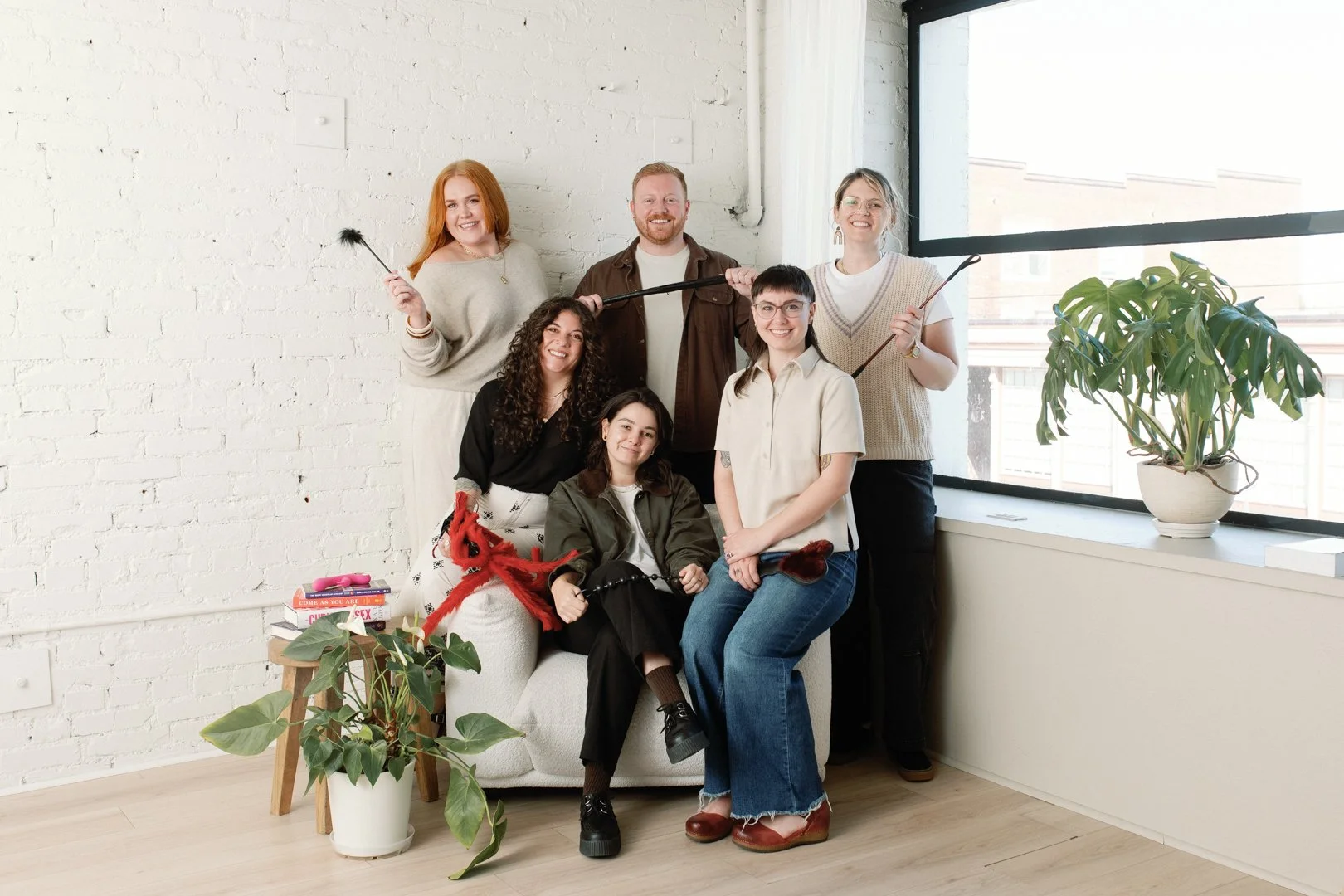 Group of seven diverse women in a bright room with a white brick wall, large window, and potted plants, posing happily with some holding overgrown cat toys or sticks.