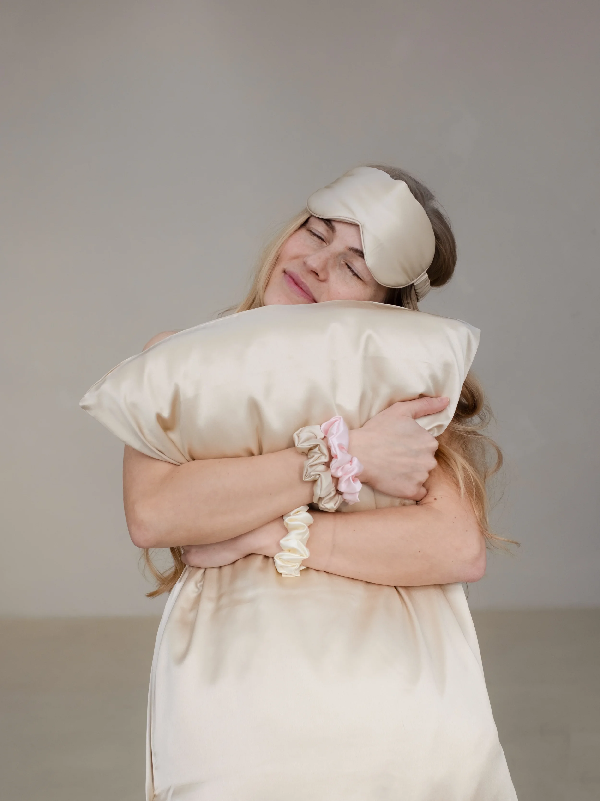 A woman with light skin and freckles hugging a satin pillow, wearing a sleep mask on her forehead, smiling with eyes closed.