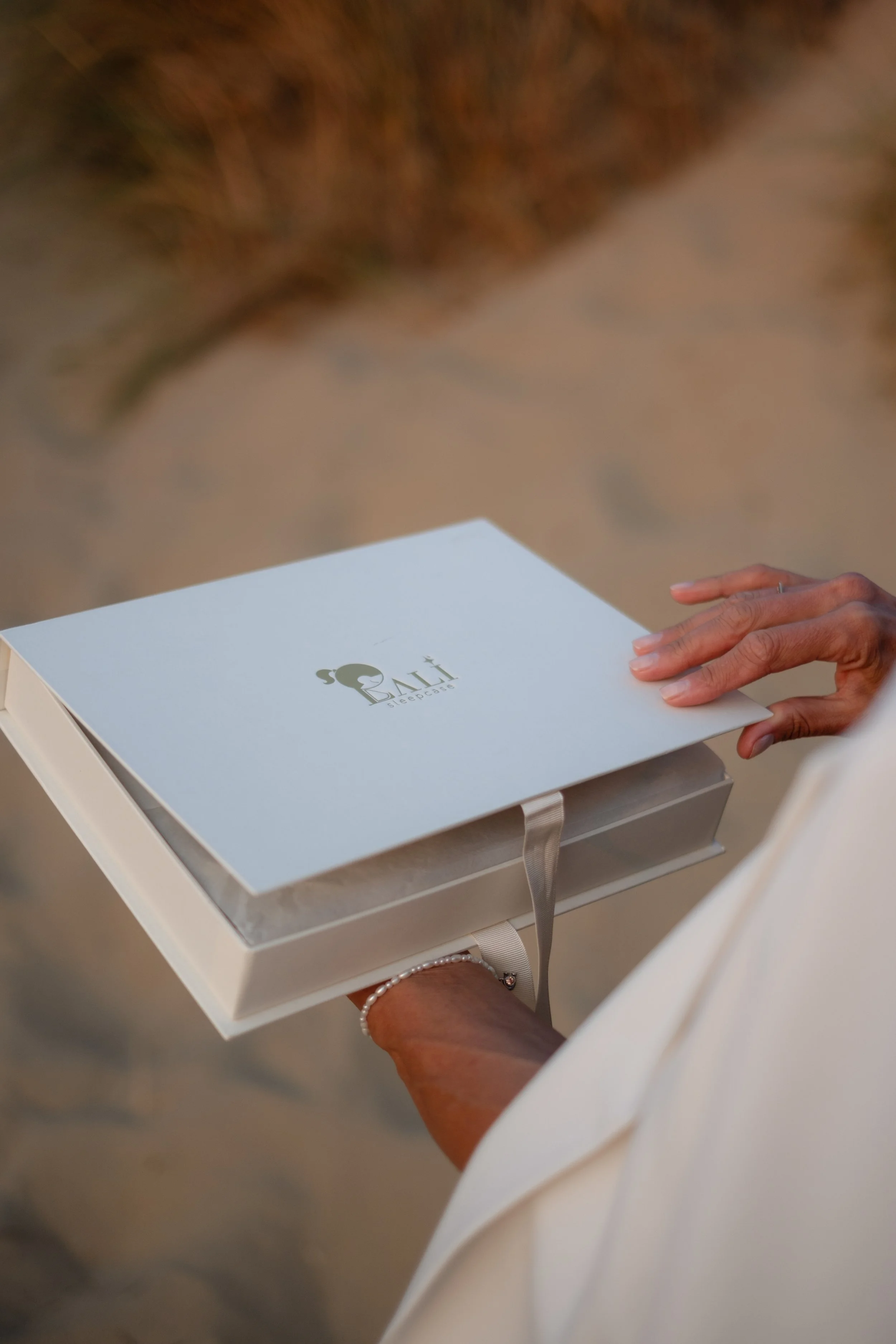 A person holding a white box with a logo that features a sculpture of two heads and the text 'BAKI gueenage'. The person is wearing a white long sleeve outfit, and a bracelet on their wrist. The background is blurred.