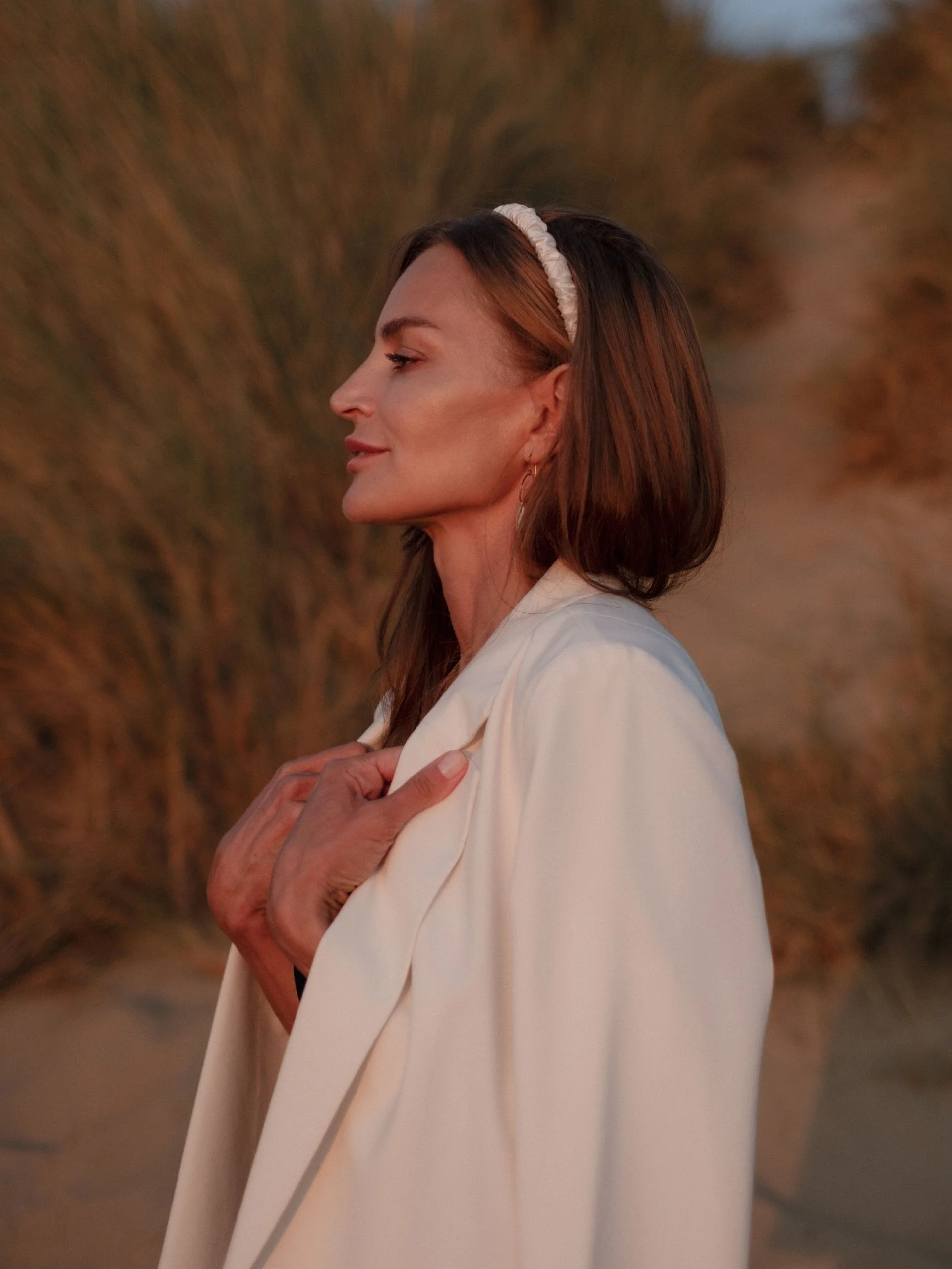 A woman with long brown hair wearing a white headband and cream-colored blazer stands outdoors. She has light makeup and multiple earrings. She is smiling slightly, with her eyes closed, and gently holds her hands over her chest, with an orange and brown blurred background.