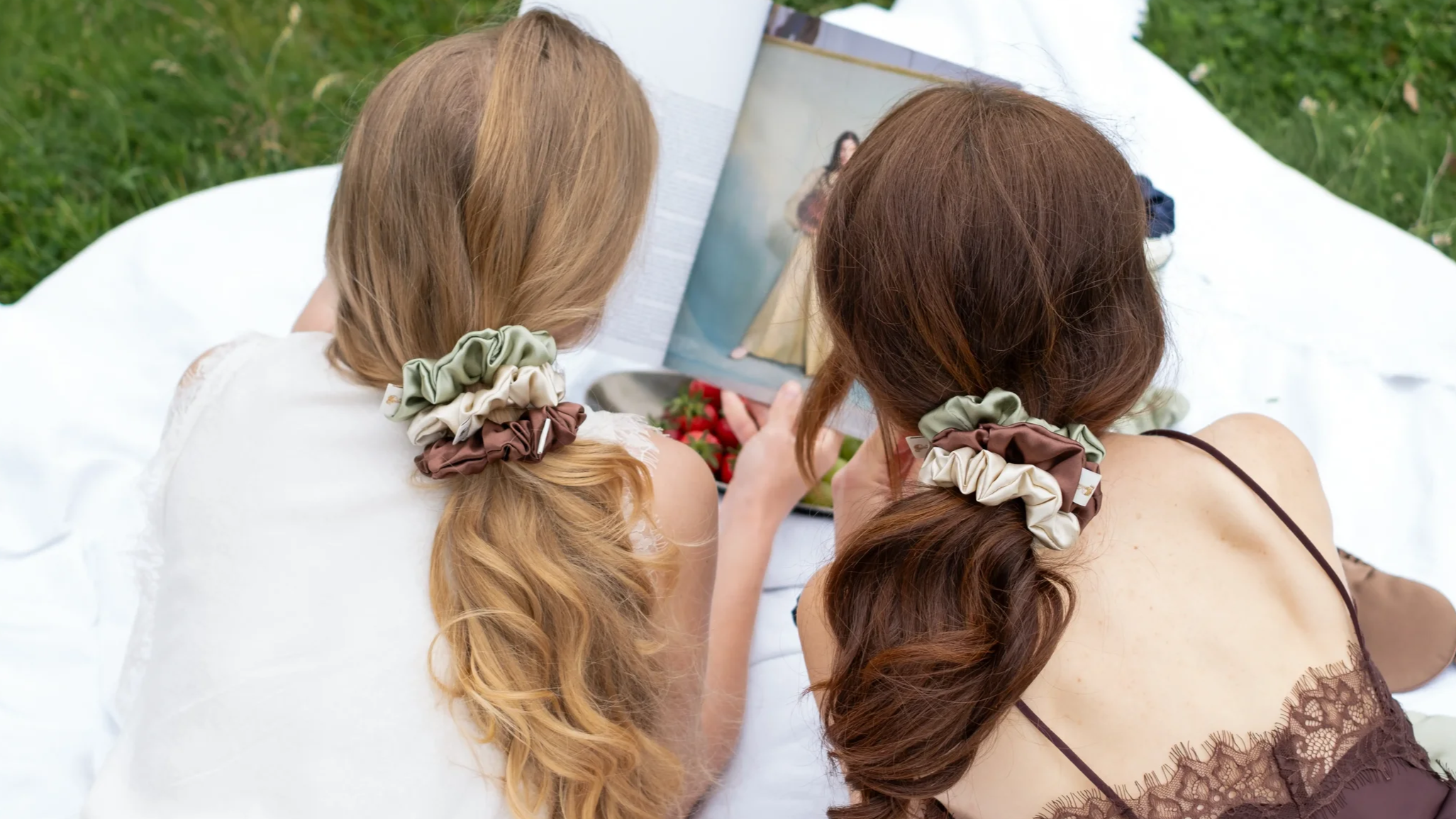Two women with matching hair scrunchies in their hair, lying on a white blanket outdoors, looking at a magazine together.