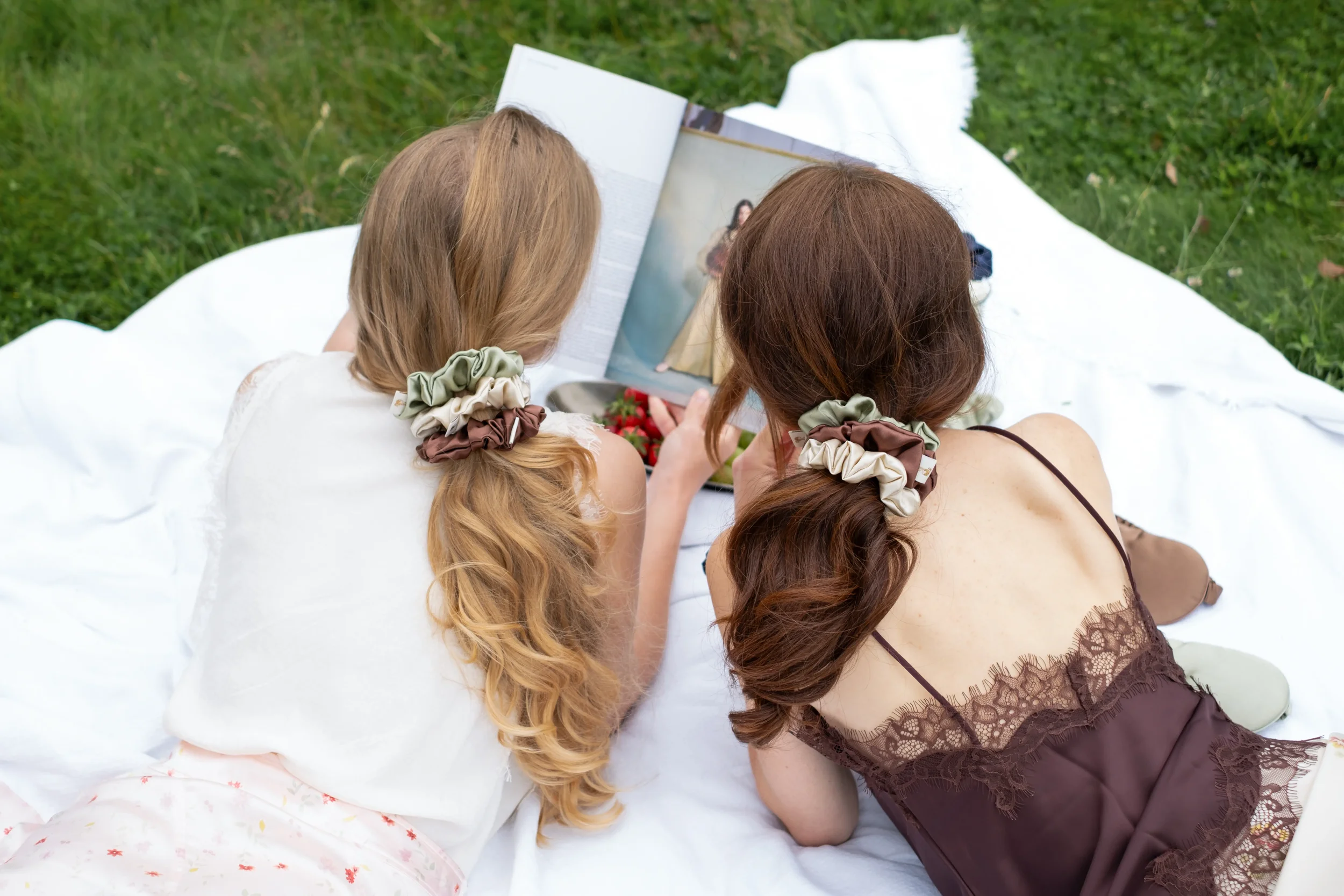 Two young women with long hair, one with blonde hair and the other with brown hair, are lying on a white blanket on the grass, looking at a magazine.