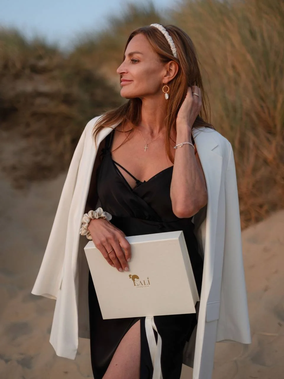 A woman with long red hair stands outdoors, smiling slightly, wearing a white blazer draped over her shoulders, a black dress, and jewelry including earrings, a necklace, and a bracelet. She holds a white box with a logo, standing on a sandy path with tall grasses in the background.