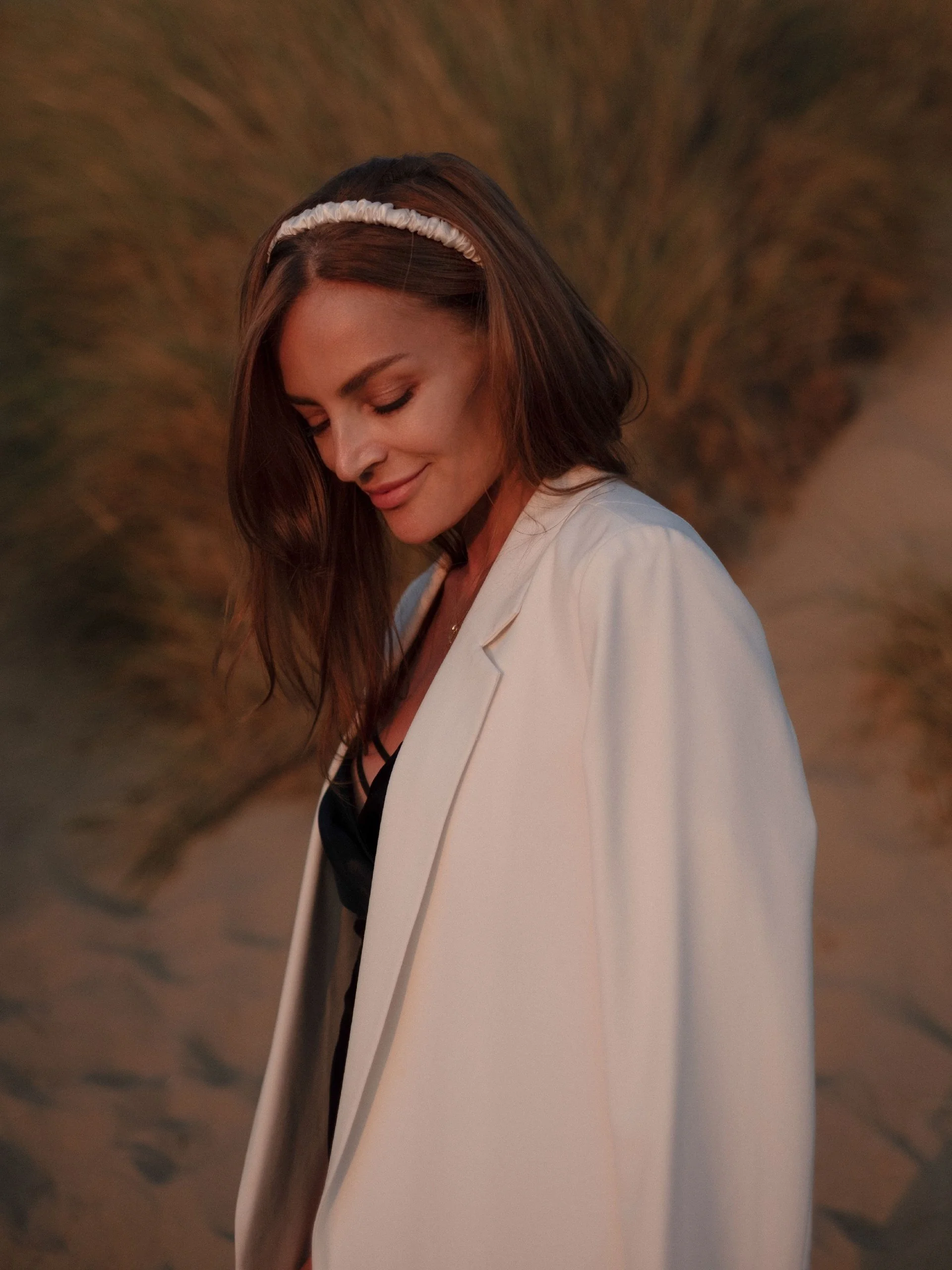 A woman with shoulder-length brown hair, wearing a white headband and a white blazer, looking down with a gentle smile, standing against a blurred, brown-toned background.