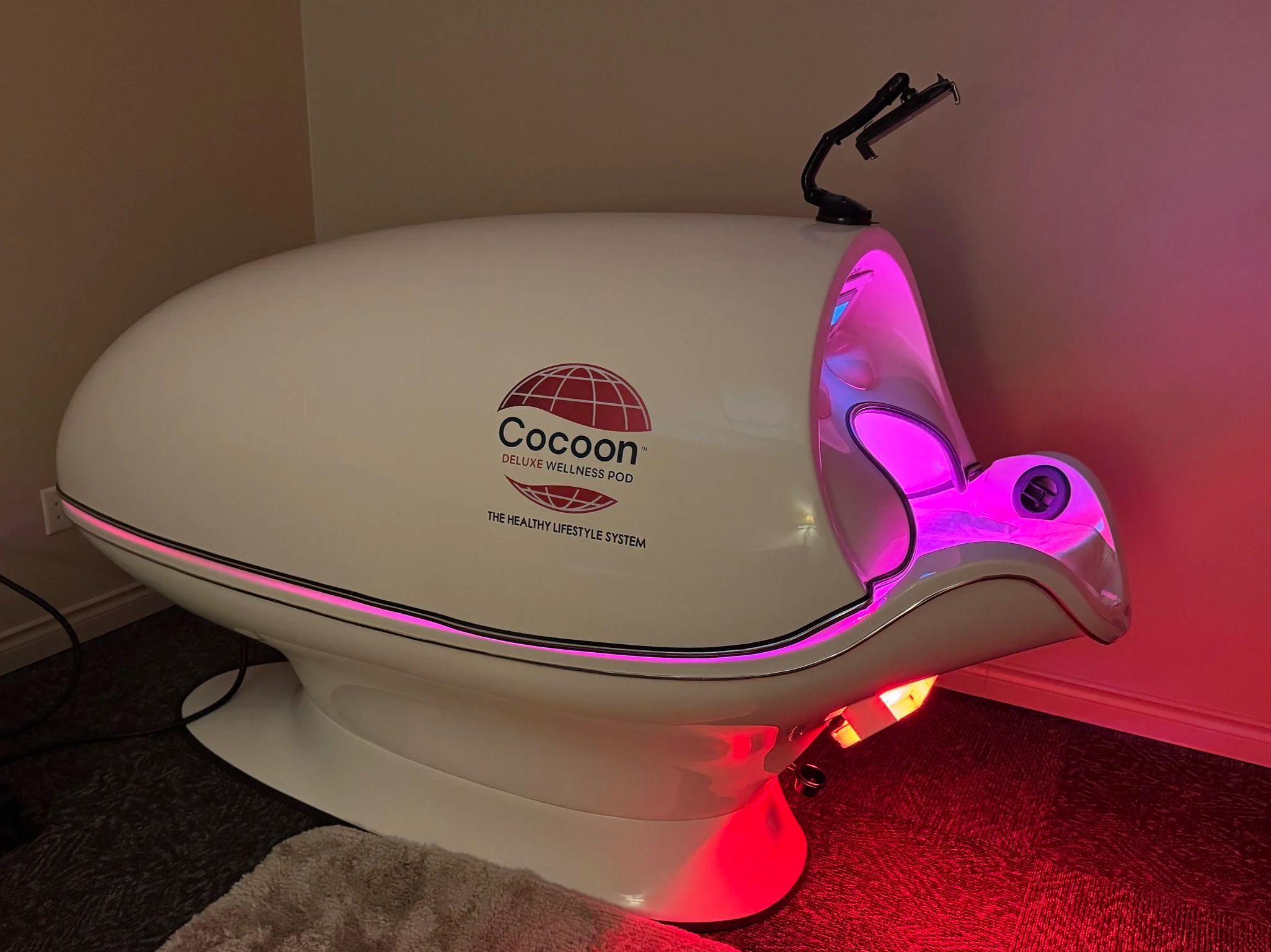 A wellness pod machine branded 'Cocoon' with pink and red LED lighting, situated on a carpeted floor in a room.