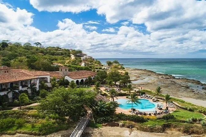 A coastal resort featuring a swimming pool, beachfront with rocky shoreline, lush greenery, and resort buildings with red tile roofs overlooking the ocean under a partly cloudy sky.