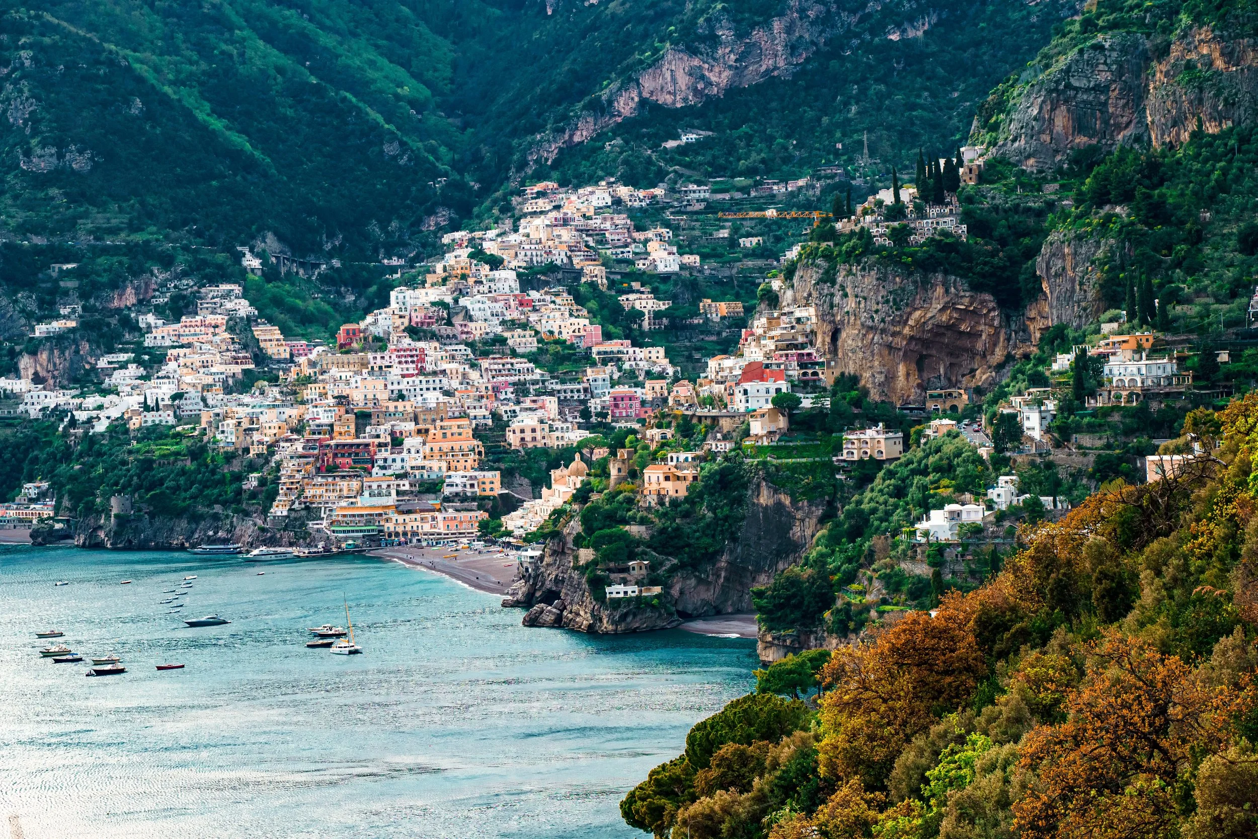 Colorful houses built on steep cliffs along the coastline in a scenic coastal town surrounded by green mountains and water with boats in the harbor.