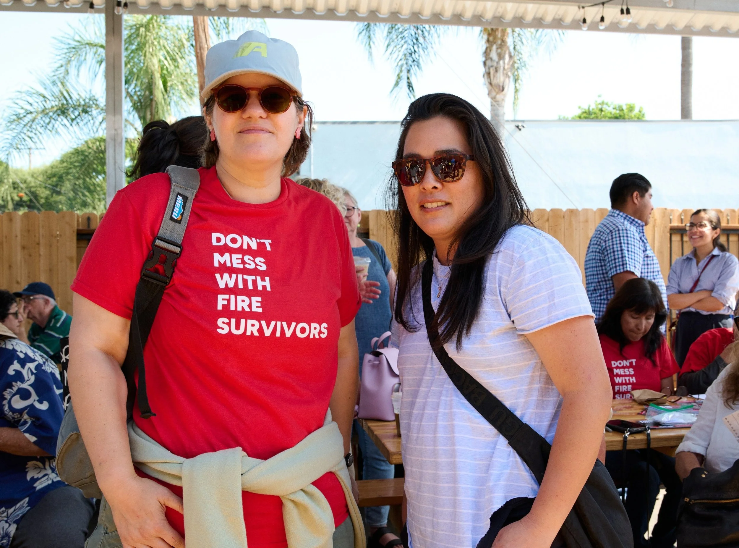 Two women outdoors, one wearing a red 'Don't Mess With Fire Survivors' t-shirt and sunglasses, both smiling