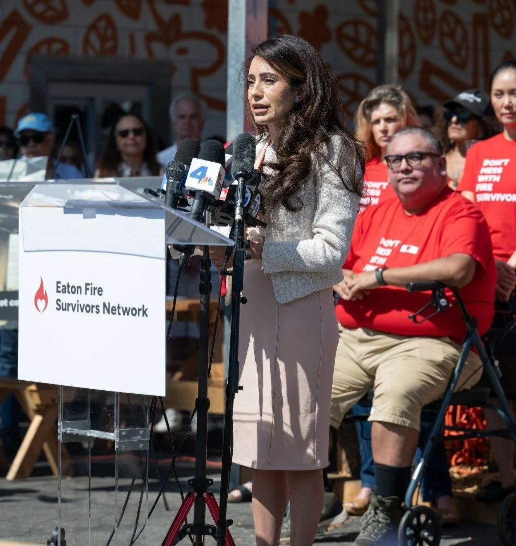 Senator Sasha Renée Pérez speaking at a podium with press microphones for the Eaton Fire Survivors Network, with survivors wearing 'Don't Mess With Fire Survivors' shirts behind her