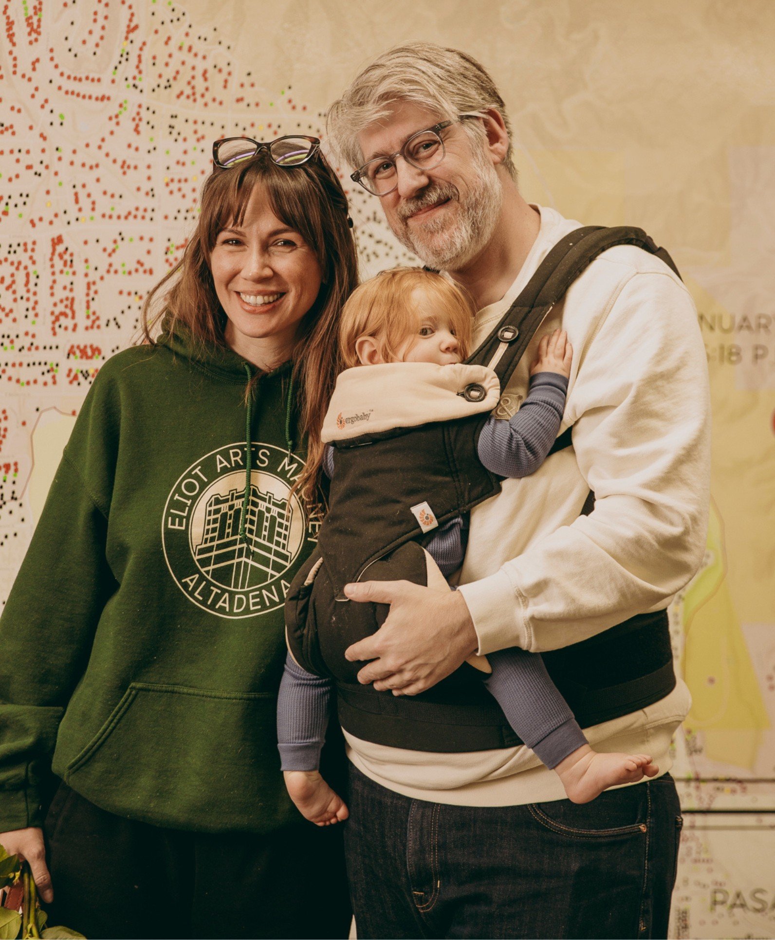 Man holding a baby in a carrier, standing next to a woman wearing an 'Eliot Arts Magnet Altadena' sweatshirt