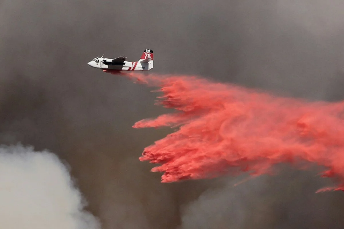 A firefighting aircraft drops bright red fire retardant from the sky against a backdrop of gray smoke clouds during aerial firefighting operations.
