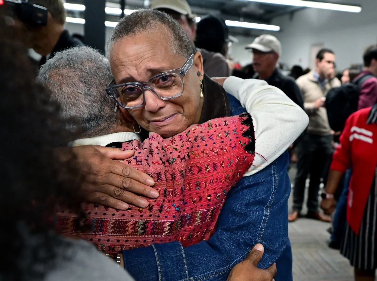 Woman with glasses embracing someone in a crowd, visibly moved