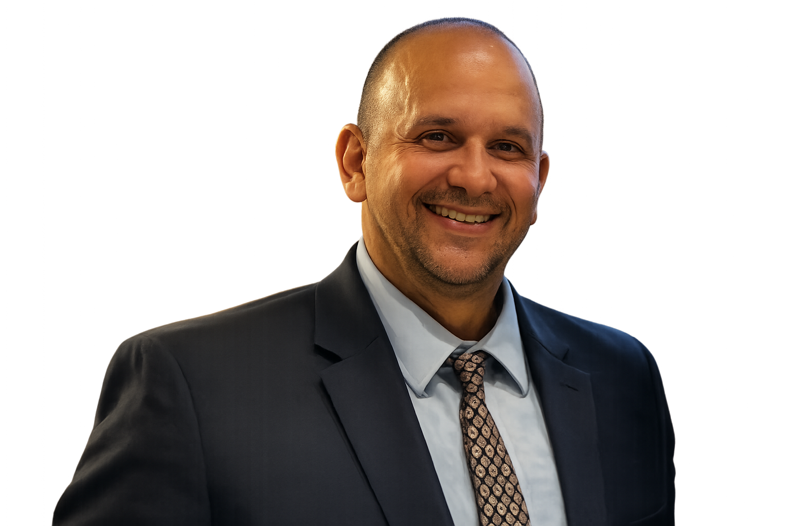A smiling man in a suit and tie standing indoors with a blurred background.